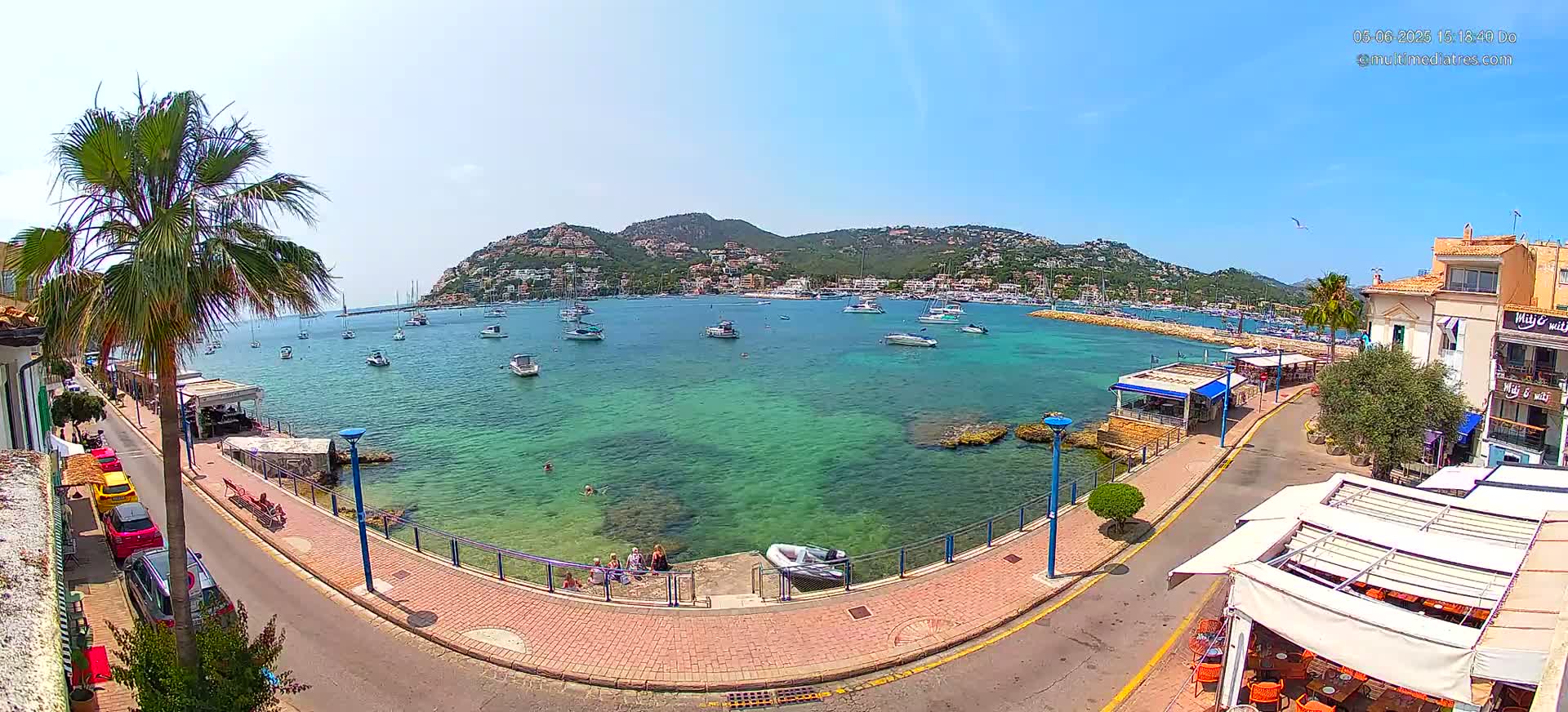 A clear, sunny day shows a calm harbor filled with boats, a town nestled against a hillside, and a paved walkway lined with restaurants and palm trees.