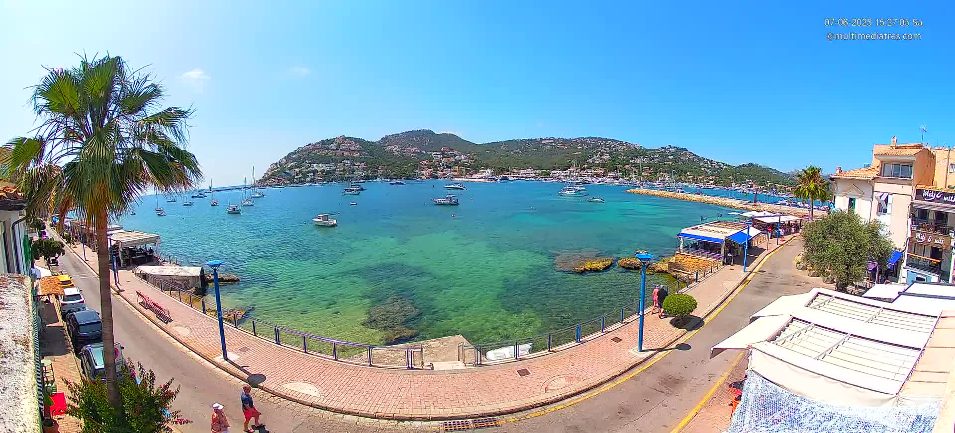 A sunny day reveals a picturesque coastal town with a calm, clear turquoise harbor filled with boats, overlooked by buildings and a hill dotted with houses.