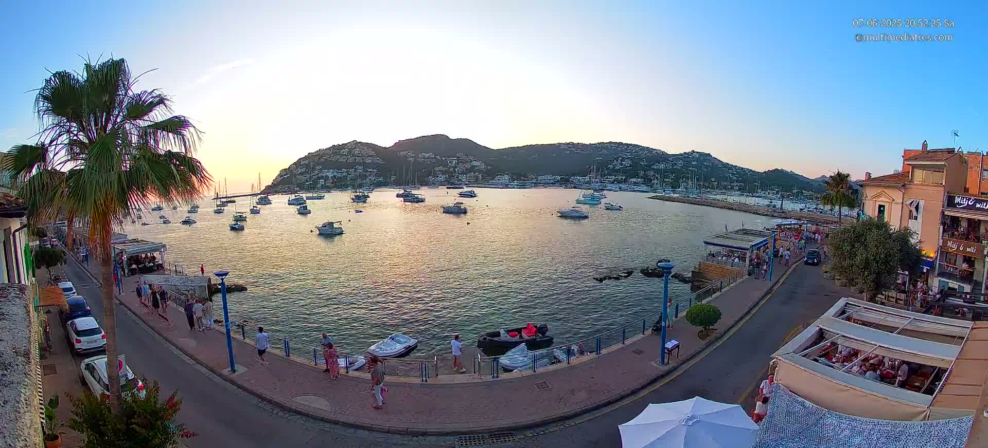 A harbor filled with boats at sunset, with people walking along a waterfront promenade next to buildings and palm trees.