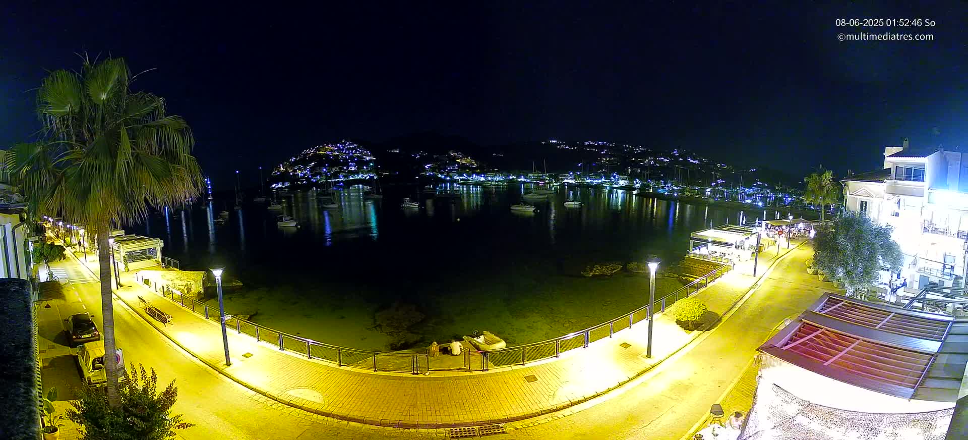 A nighttime, high-angle view shows a calm harbor with boats, illuminated buildings on a hillside, and a waterfront promenade with restaurants and palm trees under a clear, dark sky.