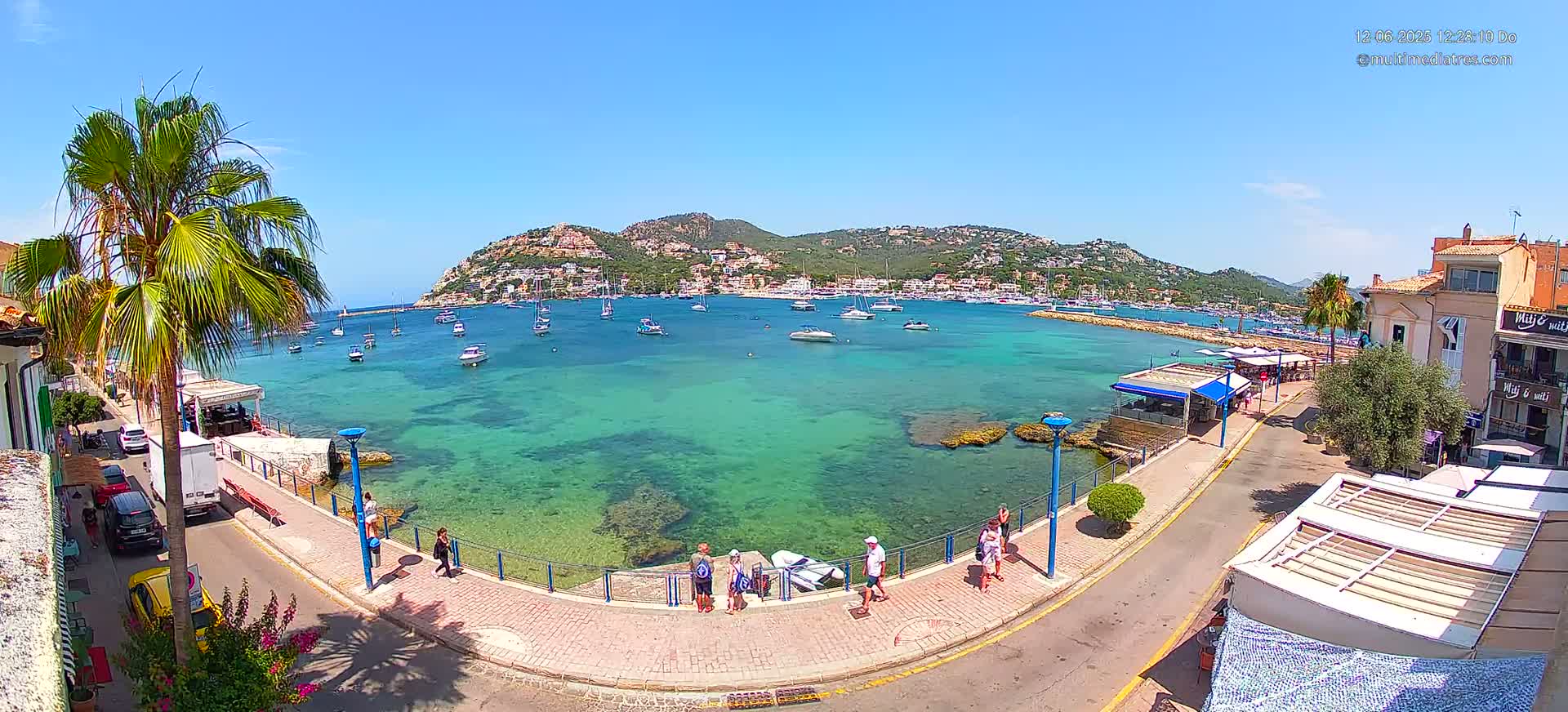 A clear, sunny day reveals a harbor filled with boats, a calm, turquoise sea, and a waterfront promenade lined with buildings and people.