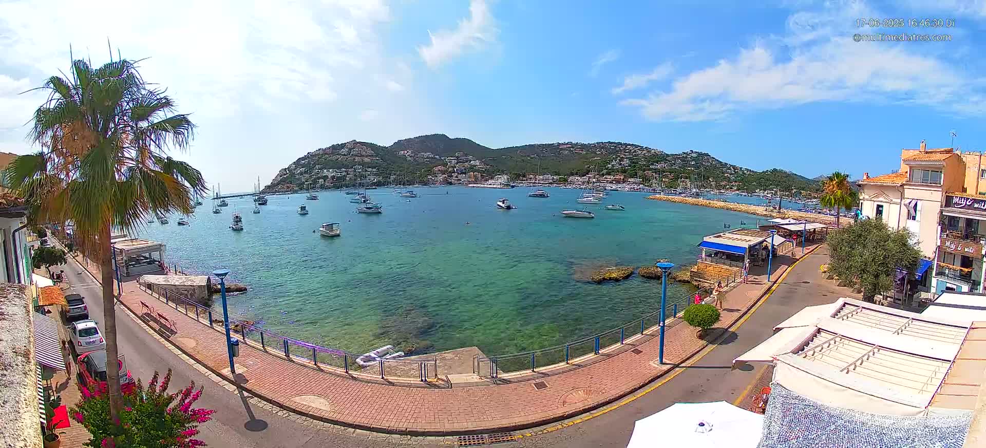 A seaside town with clear, shallow turquoise water, numerous boats in a harbor, and a hill covered in buildings in the background under a mostly sunny sky with a few clouds.
