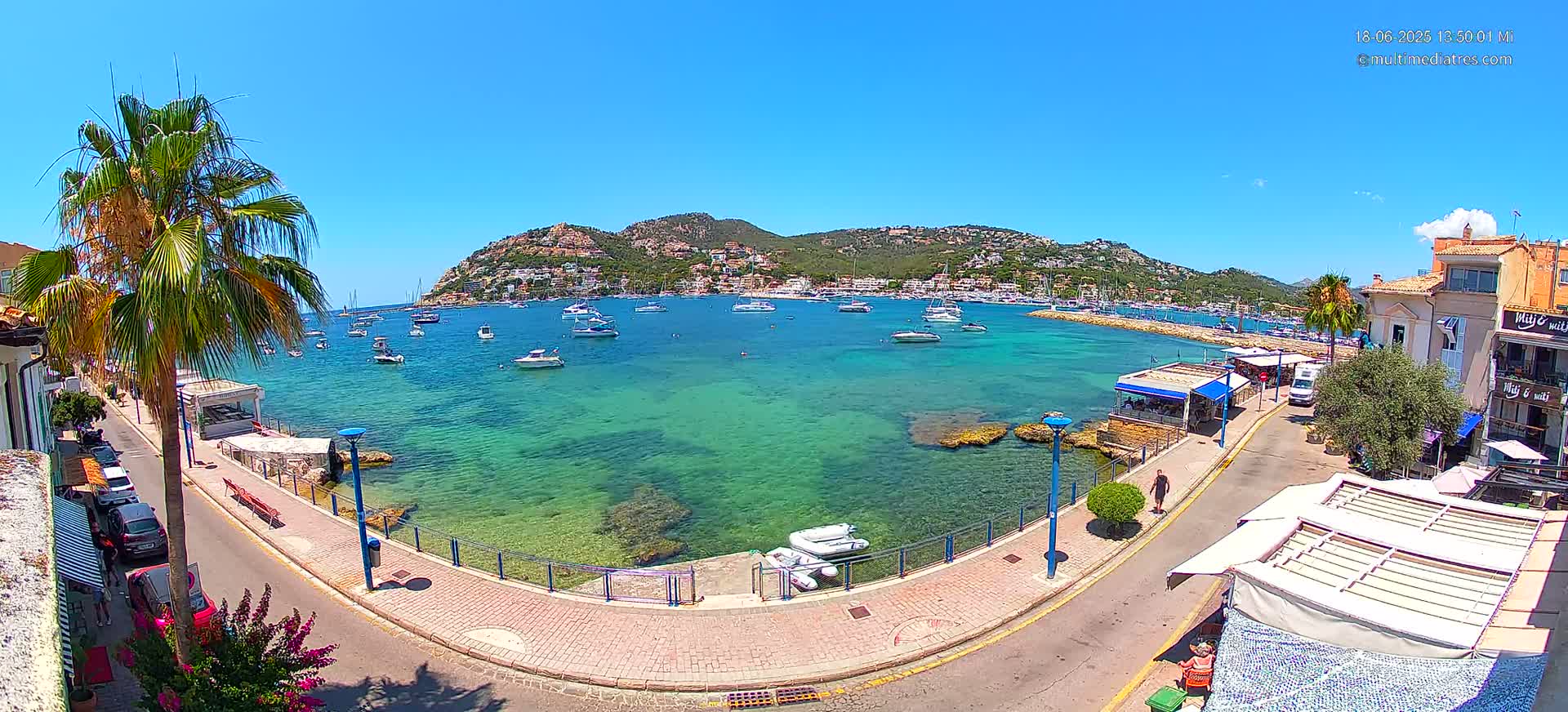 A clear, sunny day shows a calm, turquoise bay filled with numerous boats, bordered by a paved walkway, buildings, and palm trees.