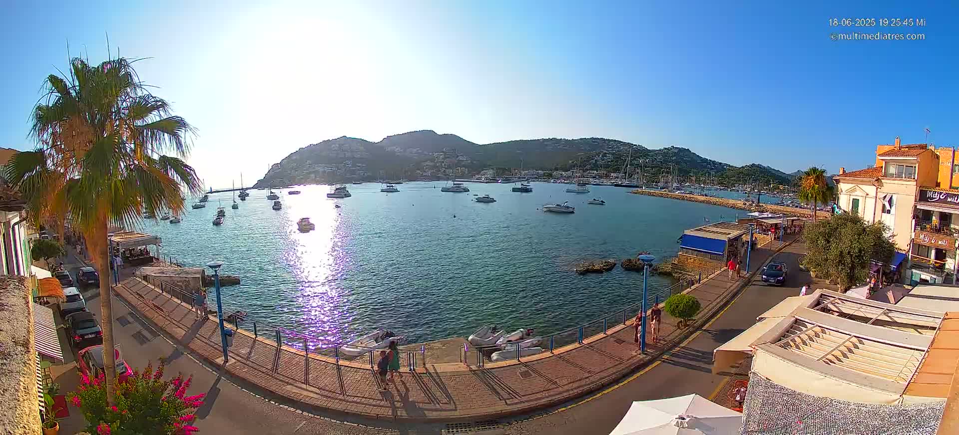 A sunny harbor scene shows numerous boats moored in calm water, a waterfront promenade with pedestrians and parked cars, and hills rising in the background.