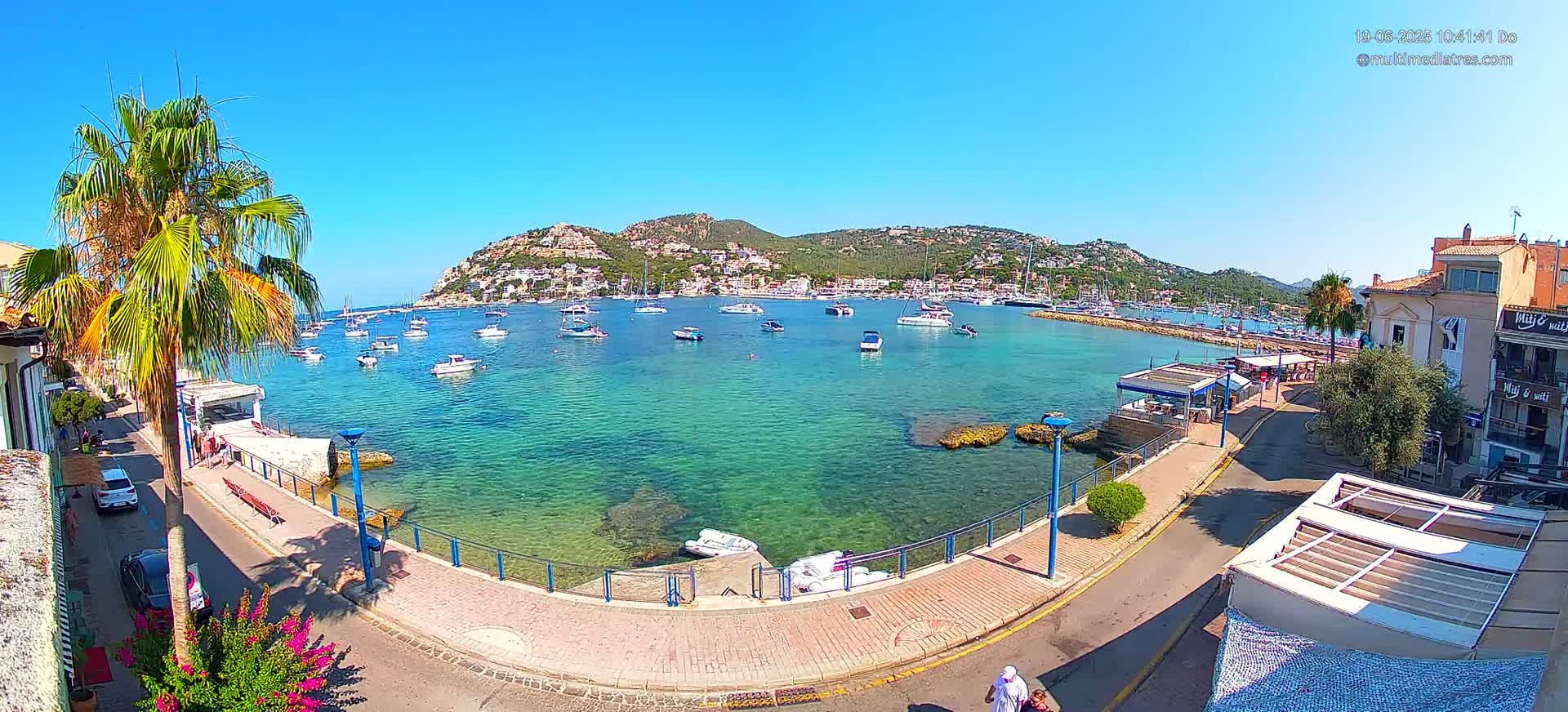 A clear, sunny day reveals a harbor filled with boats nestled beside a town situated against a backdrop of hills.
