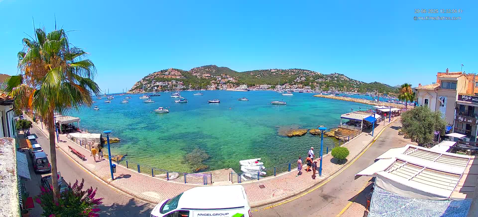 A clear, sunny day reveals a harbor filled with boats, a waterfront promenade with people walking, and a hillside town in the background.