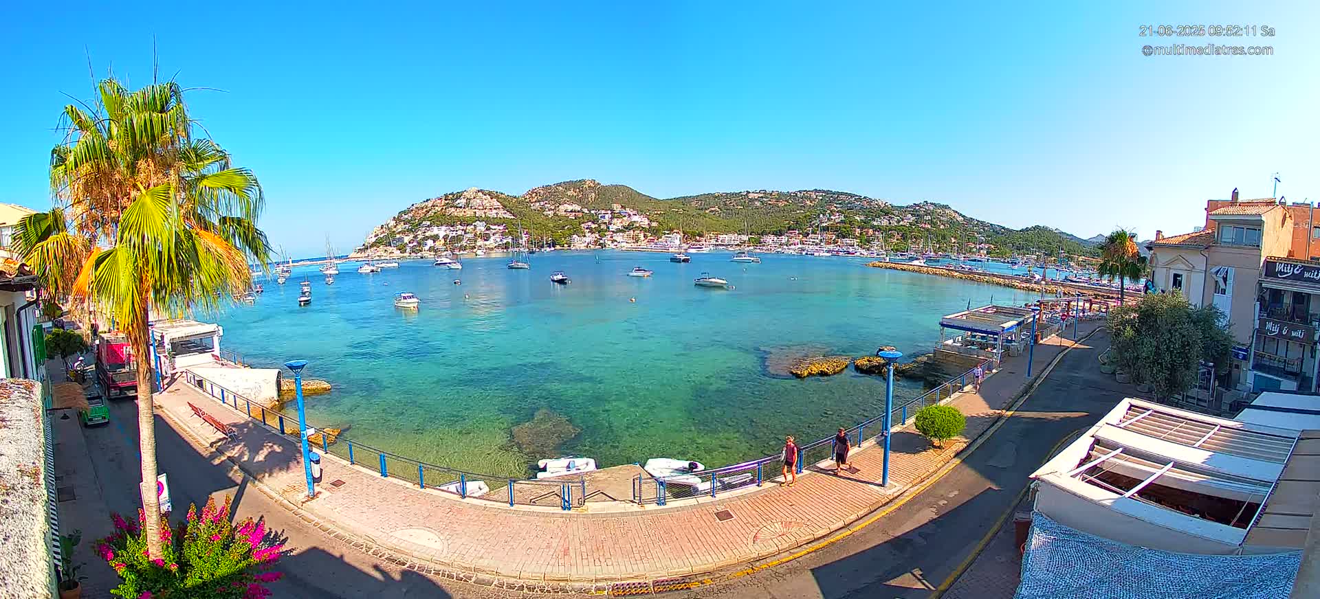 A clear sunny day shows a calm, turquoise bay filled with sailboats and motorboats, nestled between a palm-lined waterfront and a hillside town.