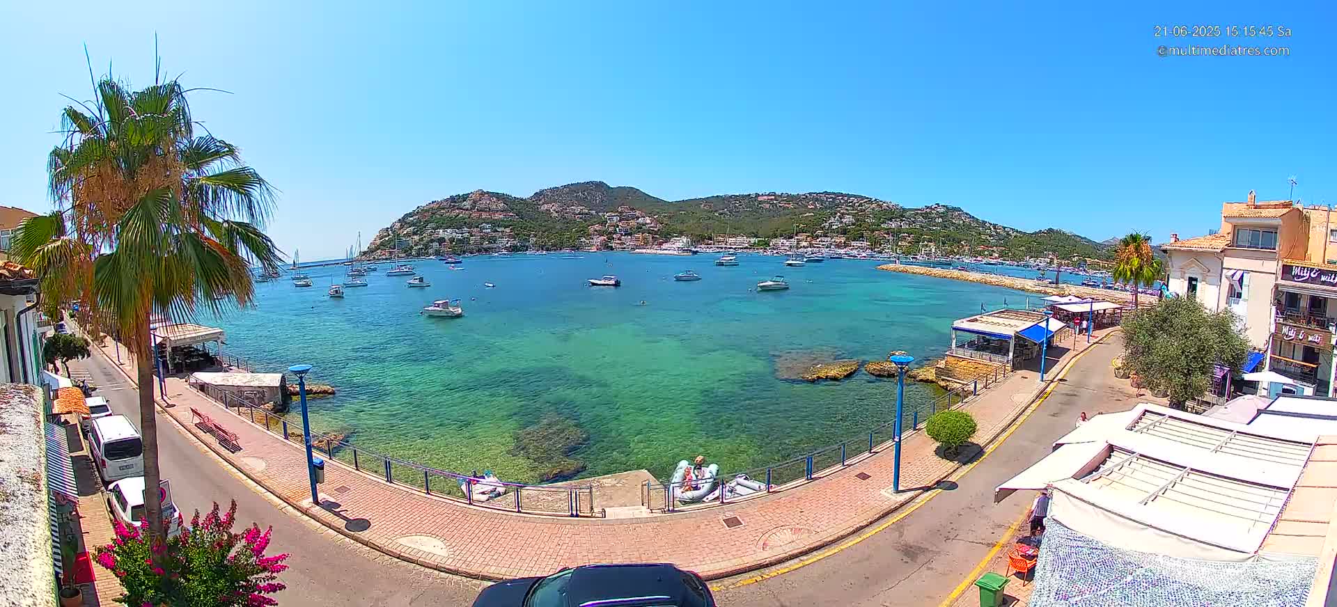 A clear sunny day shows a calm, turquoise bay with numerous boats, a waterfront promenade, and hillside buildings.