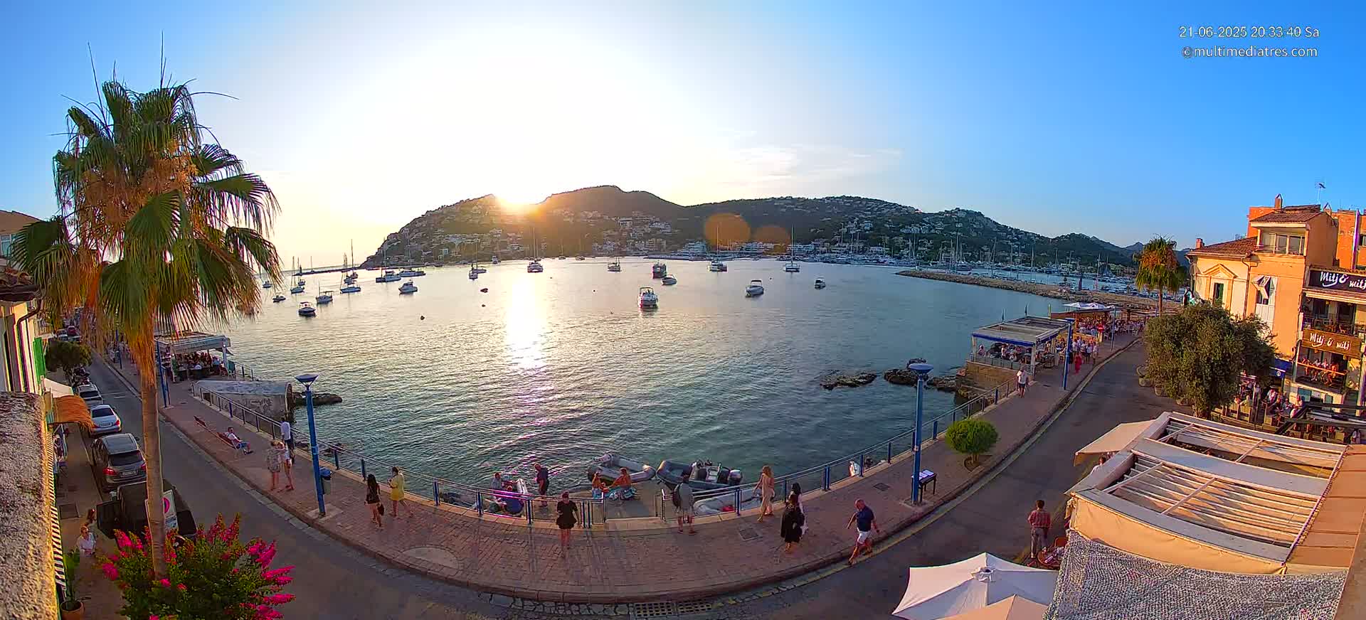 A waterfront town at sunset, with numerous boats in a calm harbor, and people strolling along a paved walkway beside restaurants and buildings.
