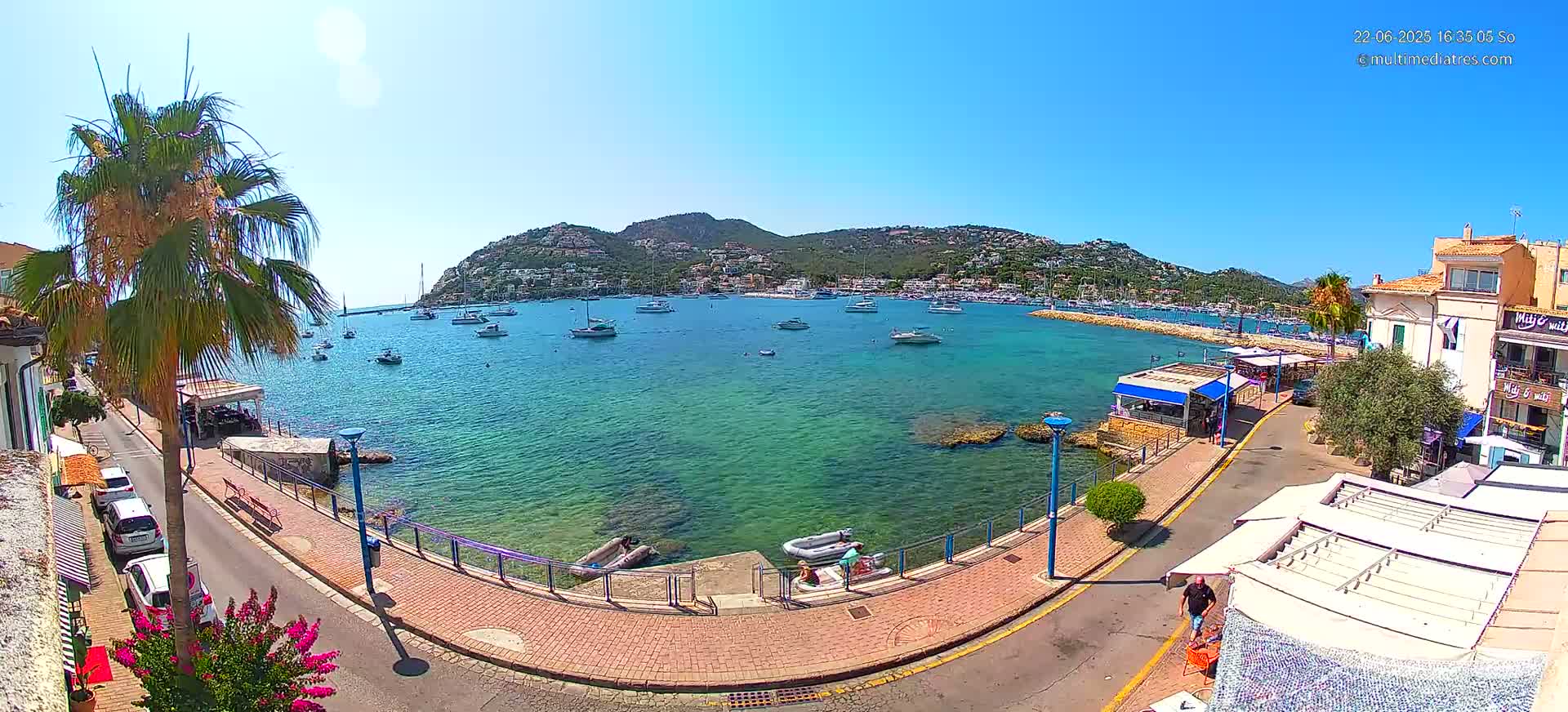 A sunny day overlooking a calm, clear, turquoise harbor filled with boats, a paved walkway along the shore lined with buildings and palm trees.