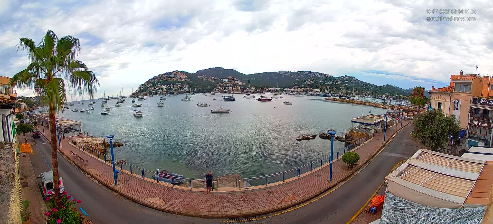 A harbor filled with numerous boats is nestled beside a town situated beneath a hill, all under a partly cloudy sky.