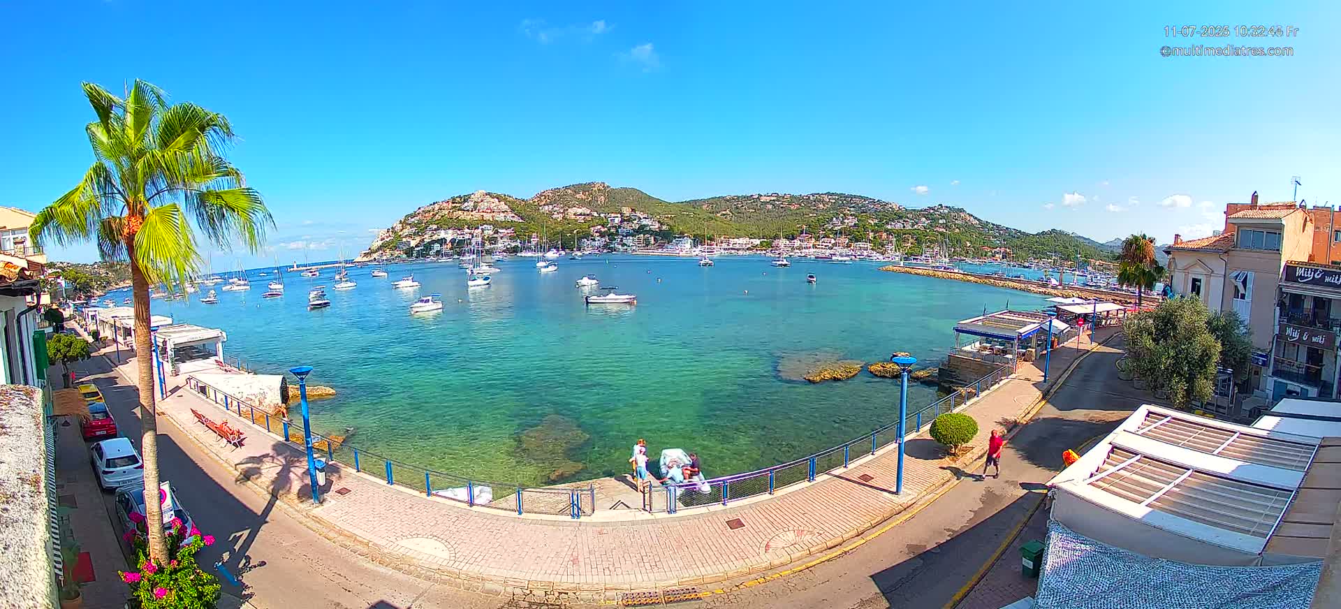 A clear, sunny day reveals a picturesque harbor filled with boats, nestled against a hillside town, with a paved walkway offering views of the turquoise water.