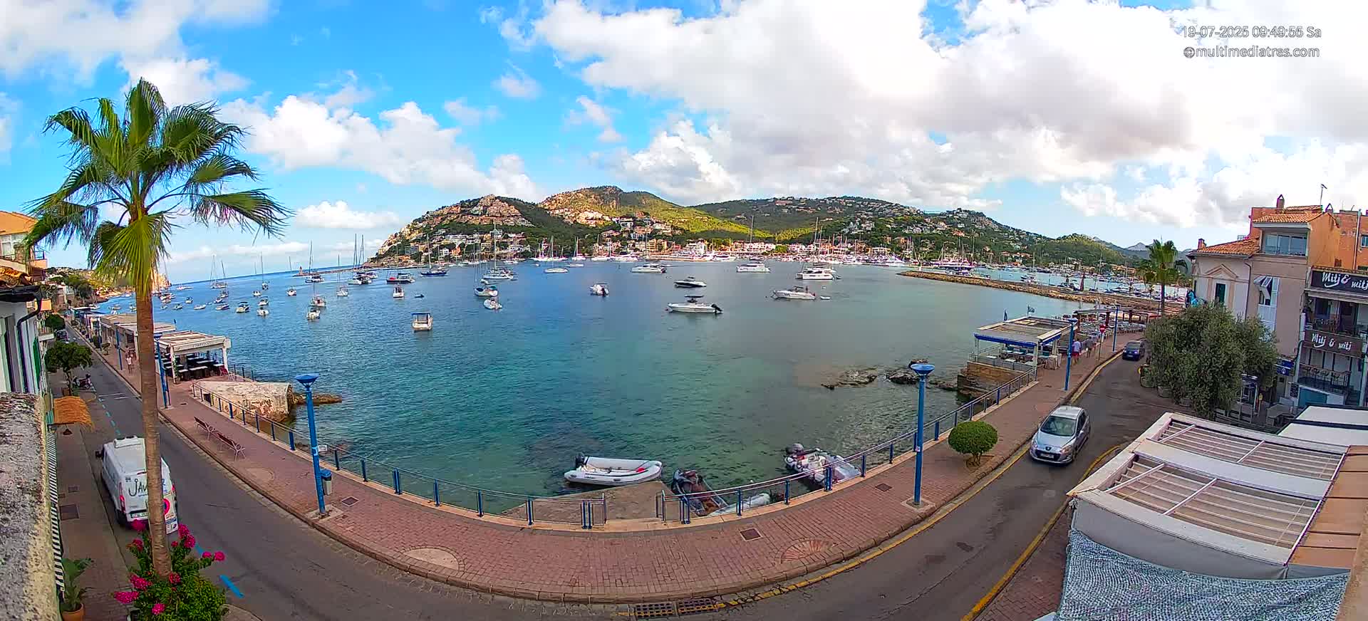 A partly cloudy day reveals a calm harbor filled with sailboats and motorboats, nestled against a hillside town under a partly cloudy sky.
