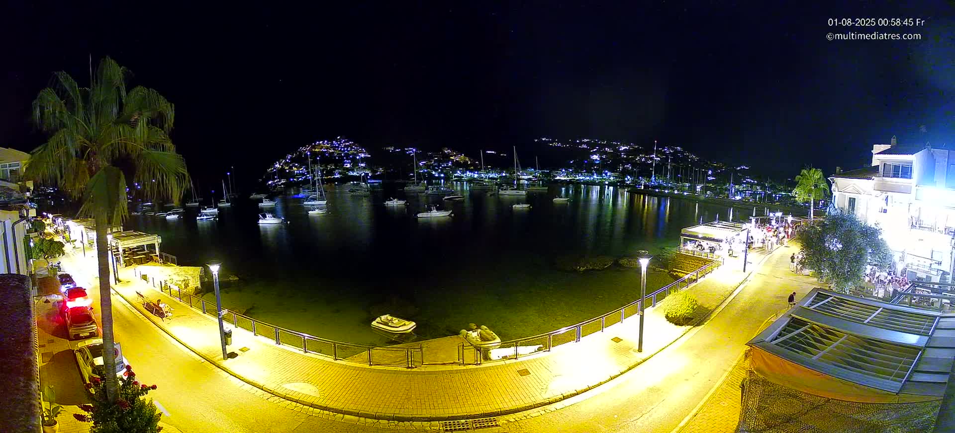 A nighttime harbor scene shows a calm body of water filled with sailboats, lined by a brightly lit walkway with people and buildings, all under a clear, dark sky.