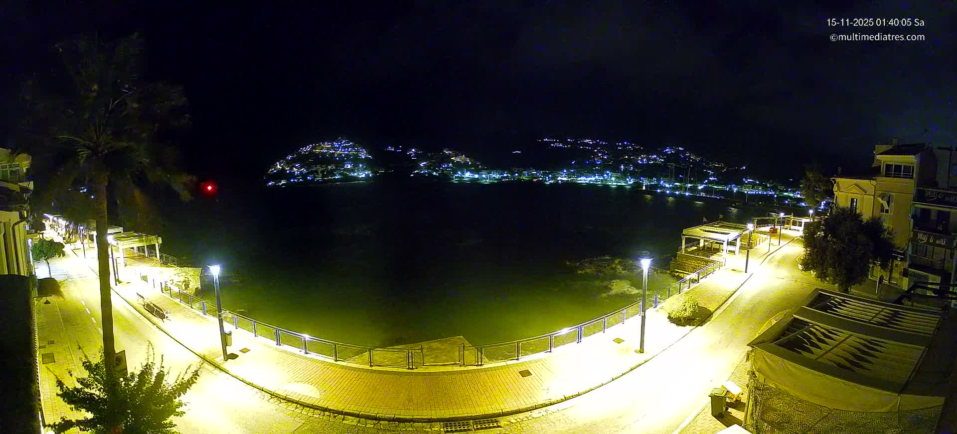 A serene night scene captures an illuminated waterfront promenade beside calm waters, with a town twinkling on distant hills under a dark, slightly cloudy sky.