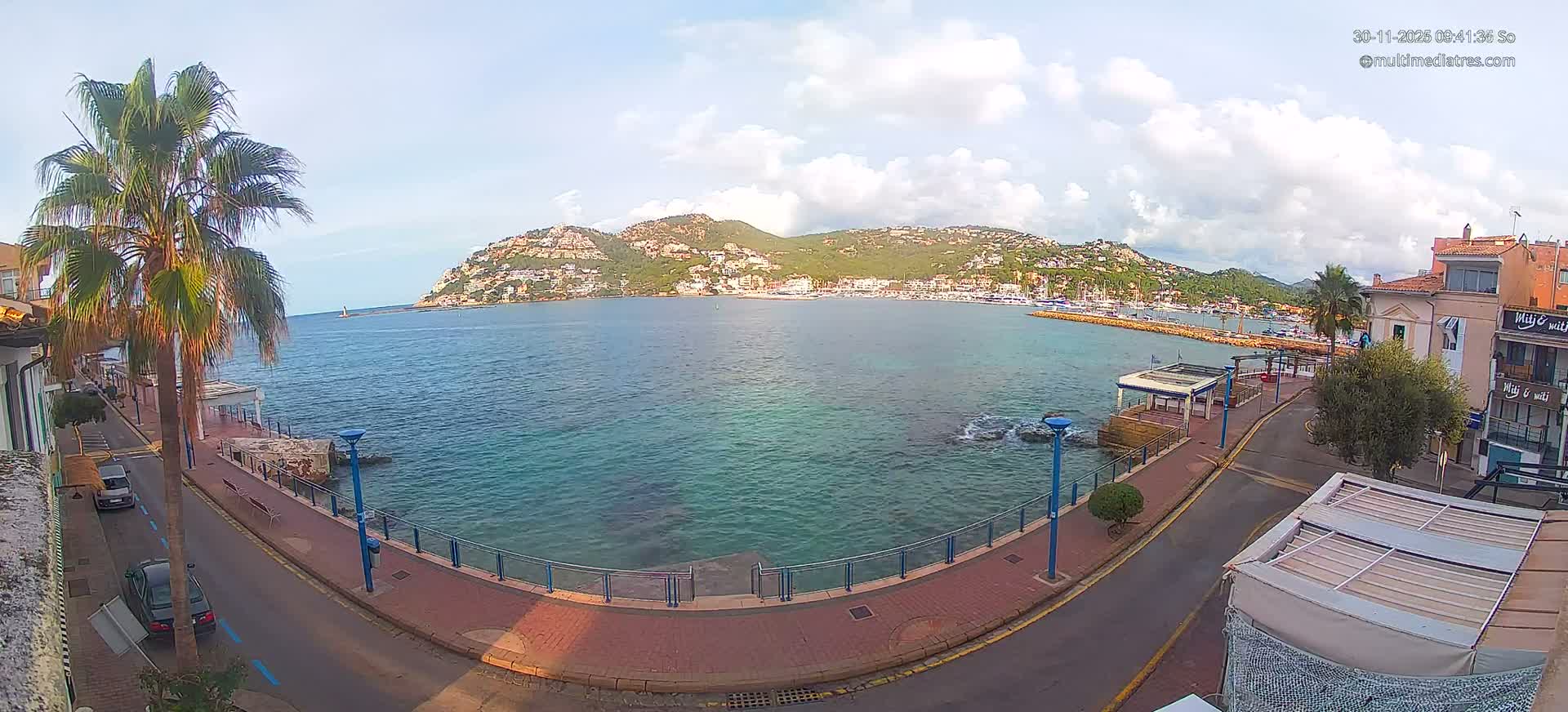 A panoramic view shows a vibrant coastal town nestled along a clear blue-green bay with a marina full of boats, a promenade, and a street with parked cars, all under a partly cloudy sky.