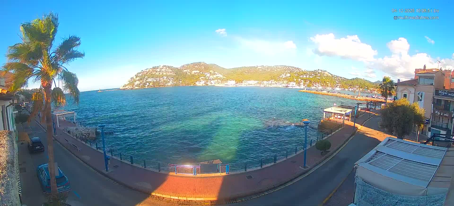 A coastal town scene on a sunny day features a clear blue bay with turquoise shallows, a curved waterfront road with buildings and palm trees, and distant green hills dotted with houses under a bright blue sky with scattered white clouds.
