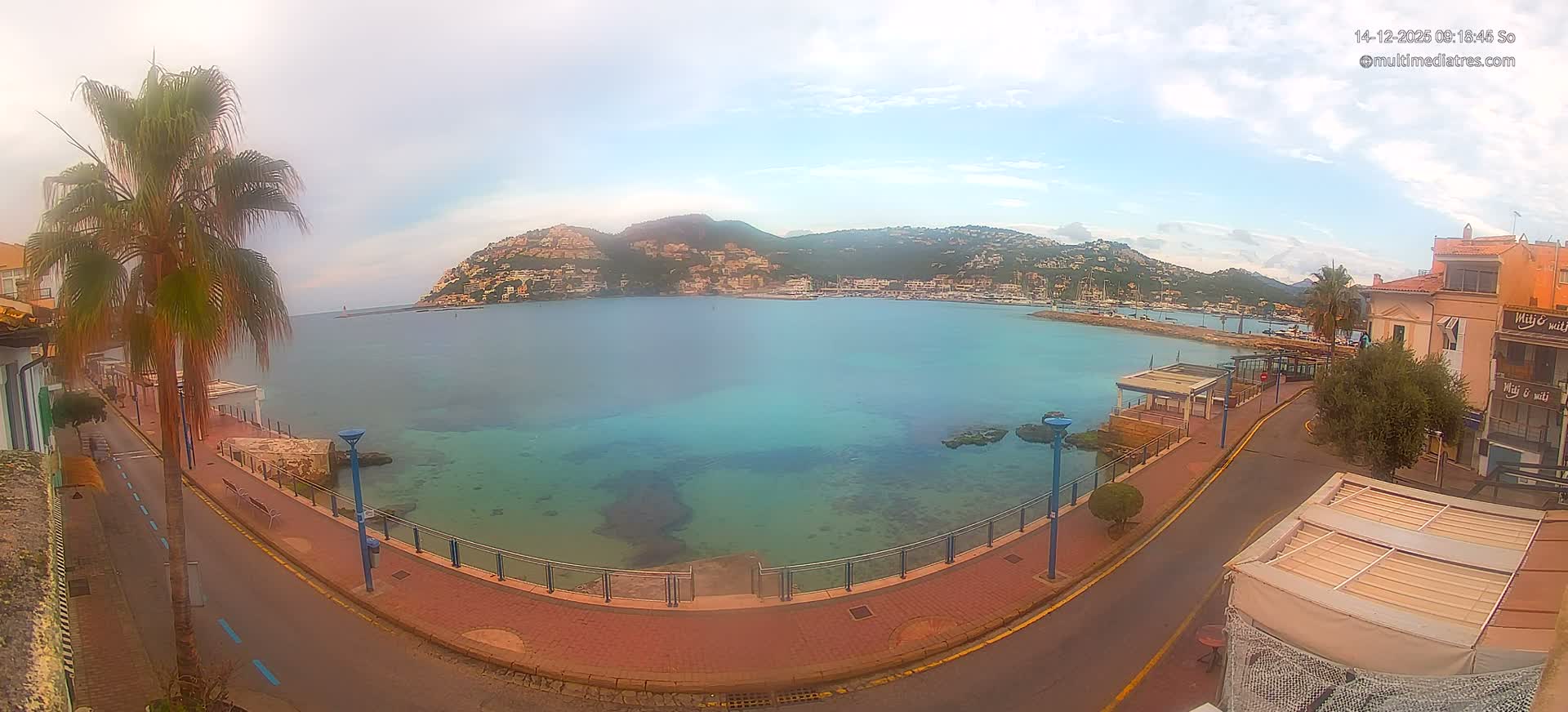 A coastal town scene on a sunny day features a clear blue bay with turquoise shallows, a curved waterfront road with buildings and palm trees, and distant green hills dotted with houses under a bright blue sky with scattered white clouds.
