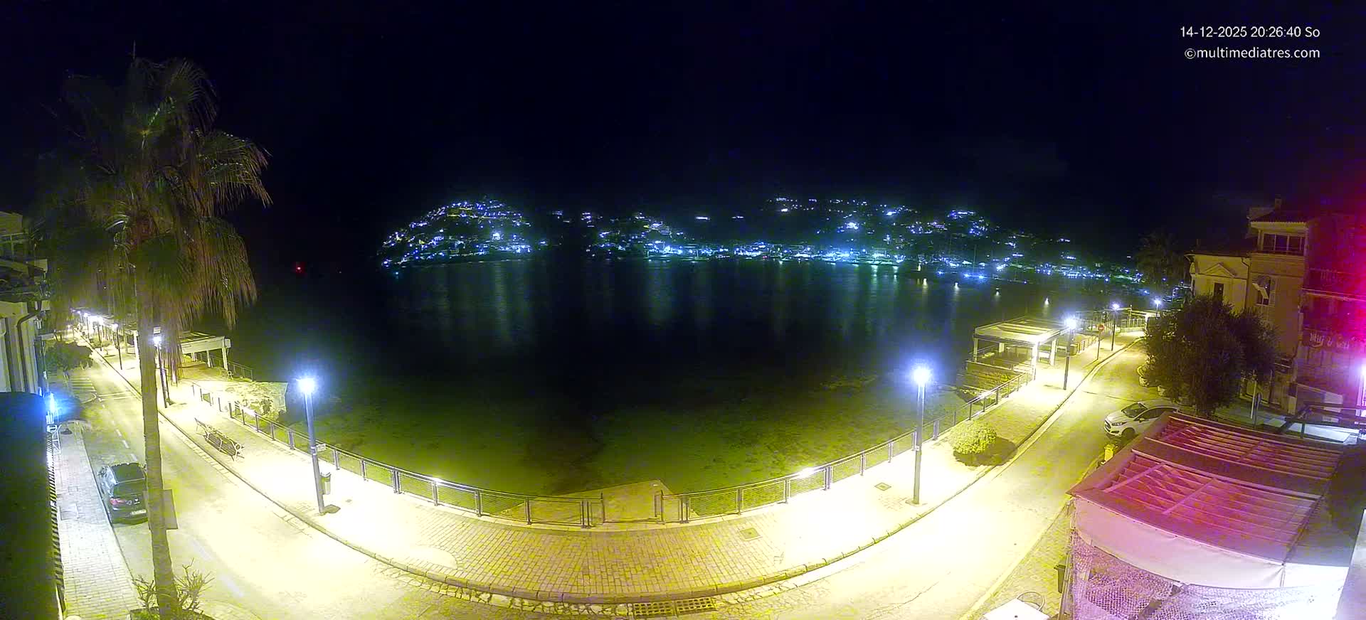 A coastal town scene on a sunny day features a clear blue bay with turquoise shallows, a curved waterfront road with buildings and palm trees, and distant green hills dotted with houses under a bright blue sky with scattered white clouds.