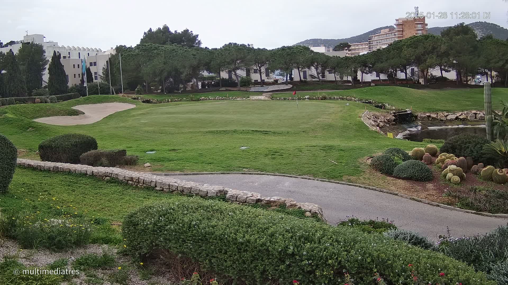 A golf course green with a sand trap, bordered by a low stone wall, under an overcast sky, with a building and numerous trees in the background.