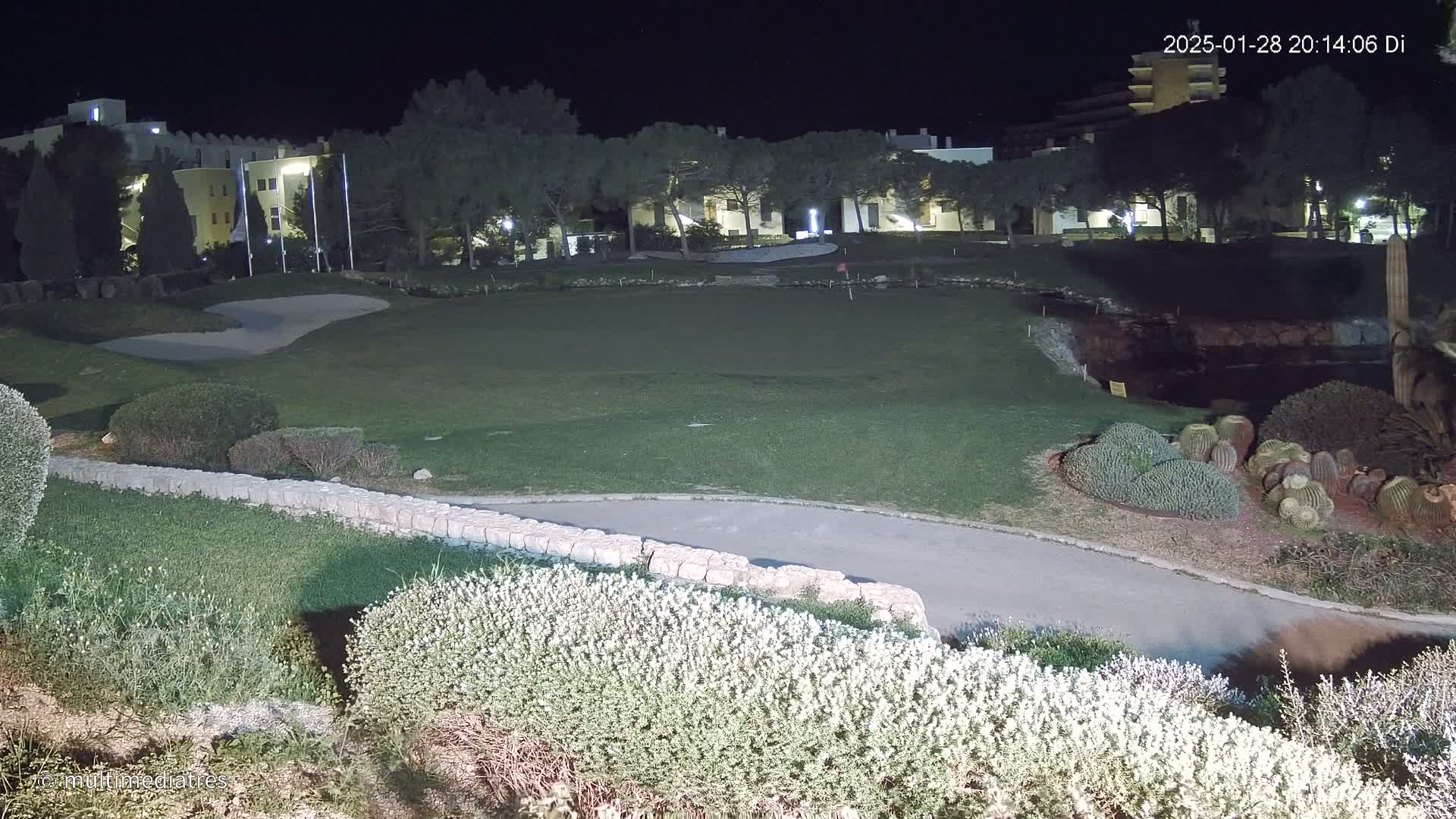 A nighttime view of a golf course with a sand trap, short grass, and low shrubbery, illuminated by artificial light, behind which are several buildings surrounded by trees.