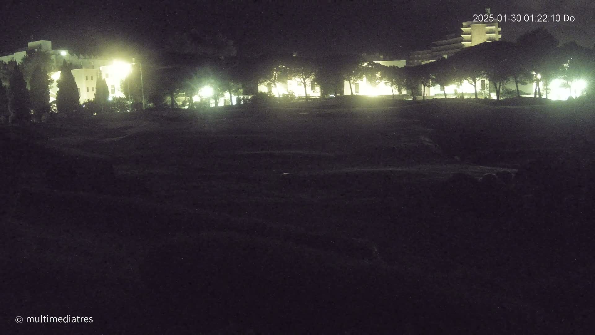 A nighttime view of illuminated buildings and trees, seen across a dark, grassy area.