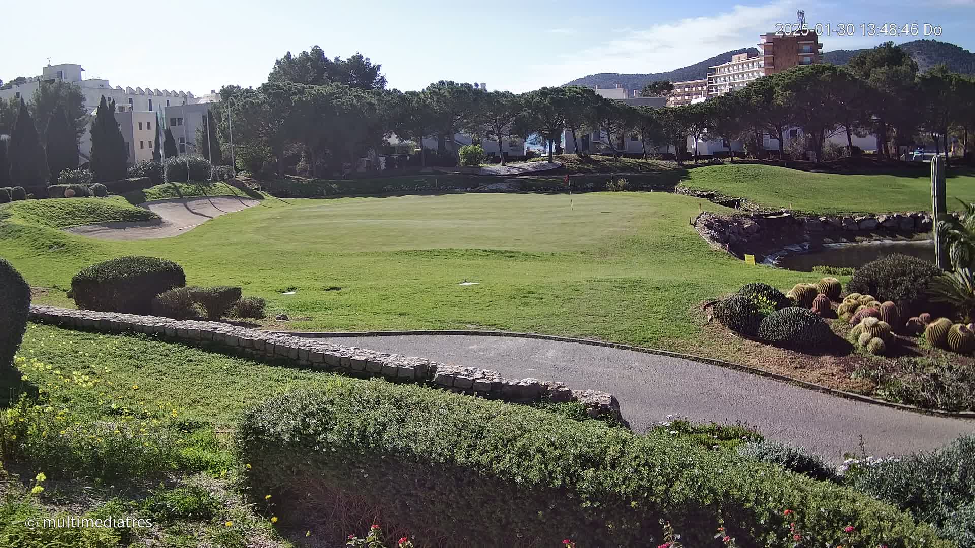 A sunny day overlooking a putting green on a golf course, with a stone path, manicured landscaping, and buildings in the background.