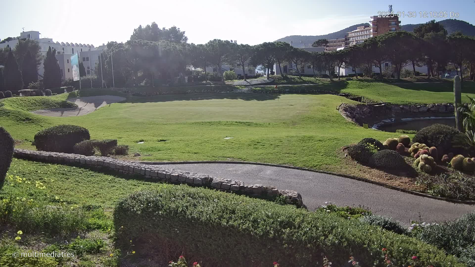 A sunny day reveals a golf course green with surrounding landscaping, a stone pathway, and buildings in the background.