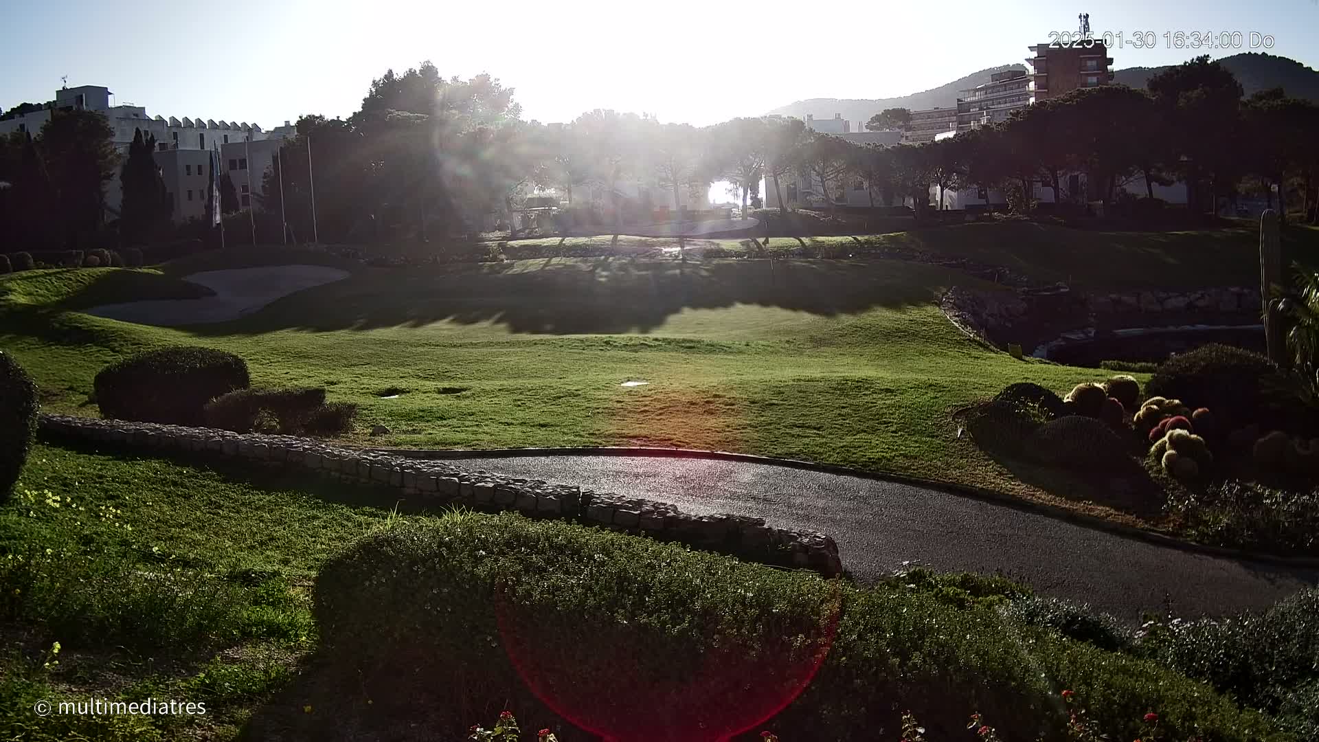 A sunny, partly-shadowed golf course with a paved path winds through manicured lawns and shrubbery,  backed by buildings and trees.