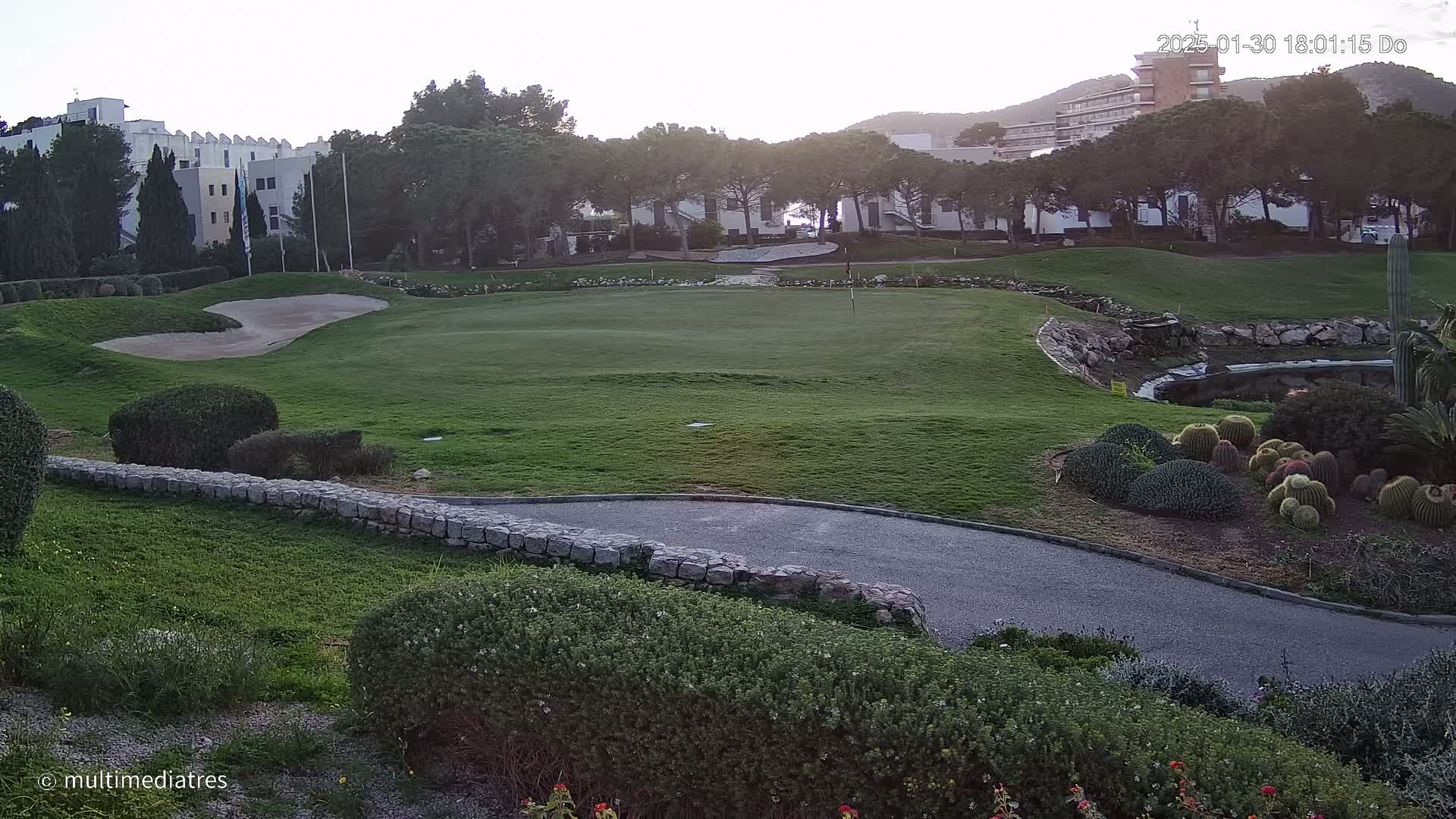 A green golf course with a sand trap, bordered by shrubs, a stone wall, and a paved path, is situated near buildings and trees under a twilight sky.