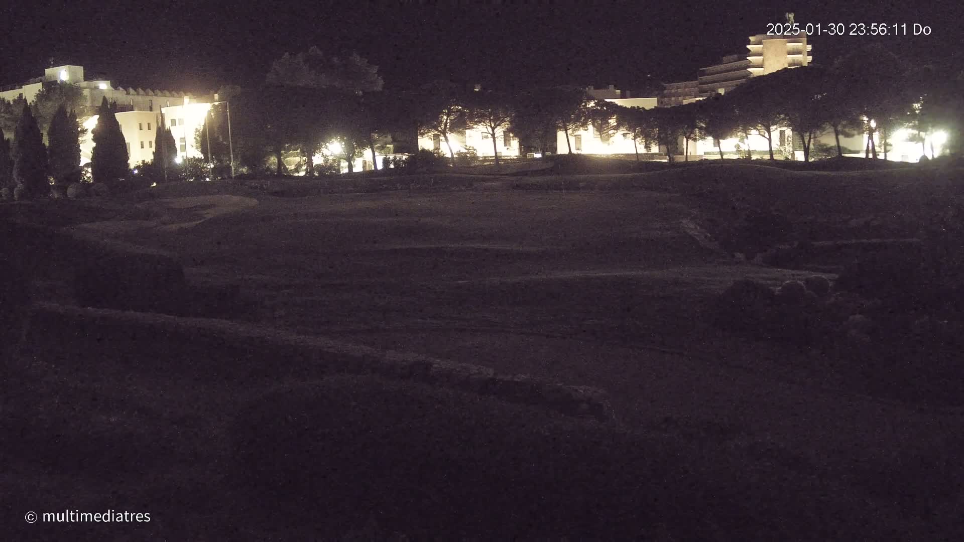 At night, a dark, possibly excavated, area is seen in front of a row of lit buildings and trees.