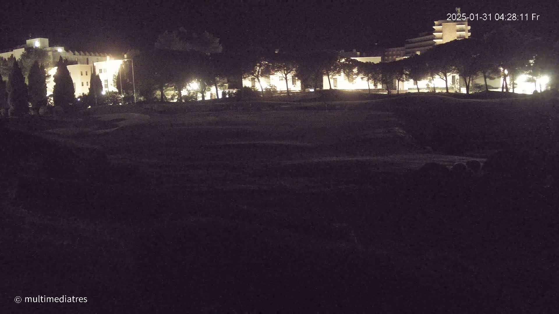 At night, illuminated buildings are partially visible behind a dark, grassy area.