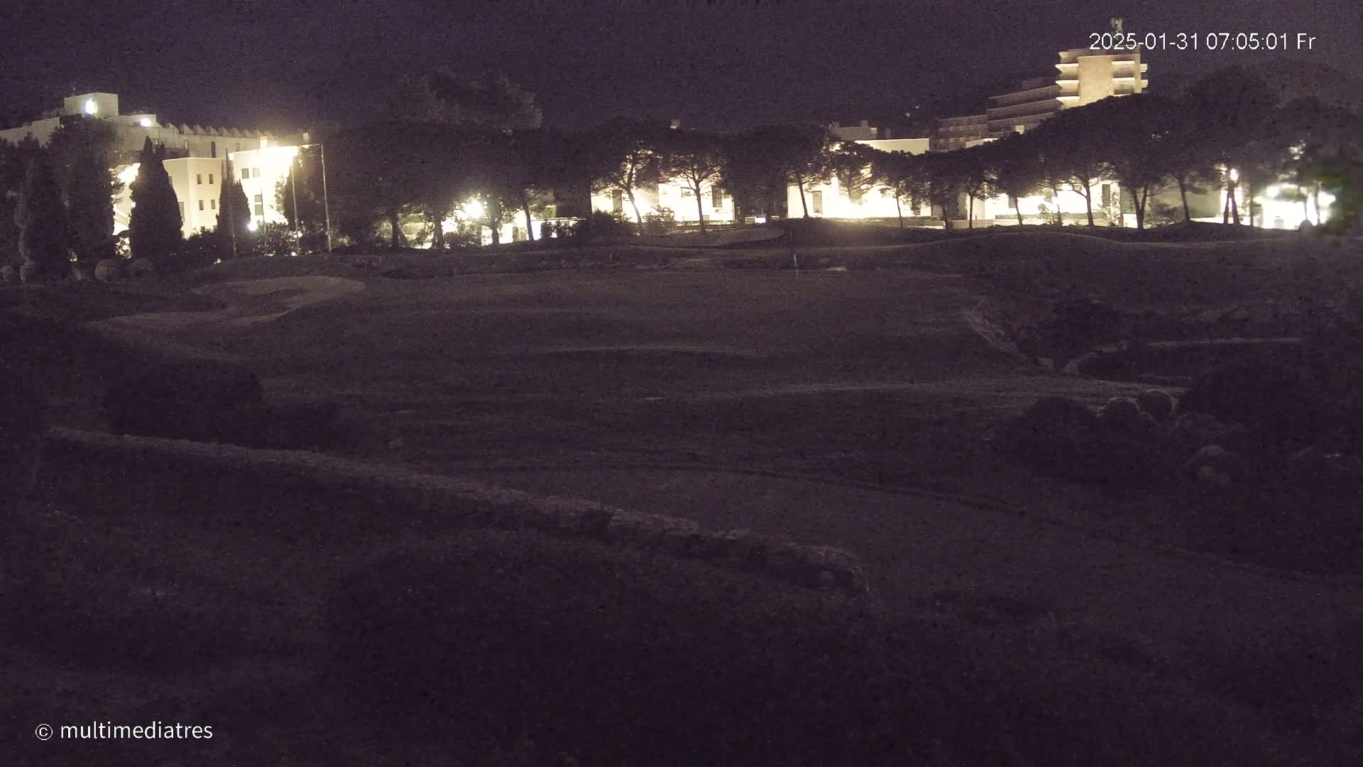 At night, a dark, grassy area with subtle undulations is illuminated by the lights of buildings and trees in the background.
