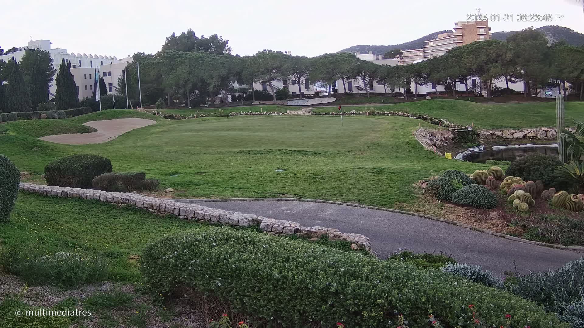 A golf course green with a sand trap, bordered by low stone walls and landscaping that includes cacti and shrubs, is situated under an overcast sky near buildings and trees.