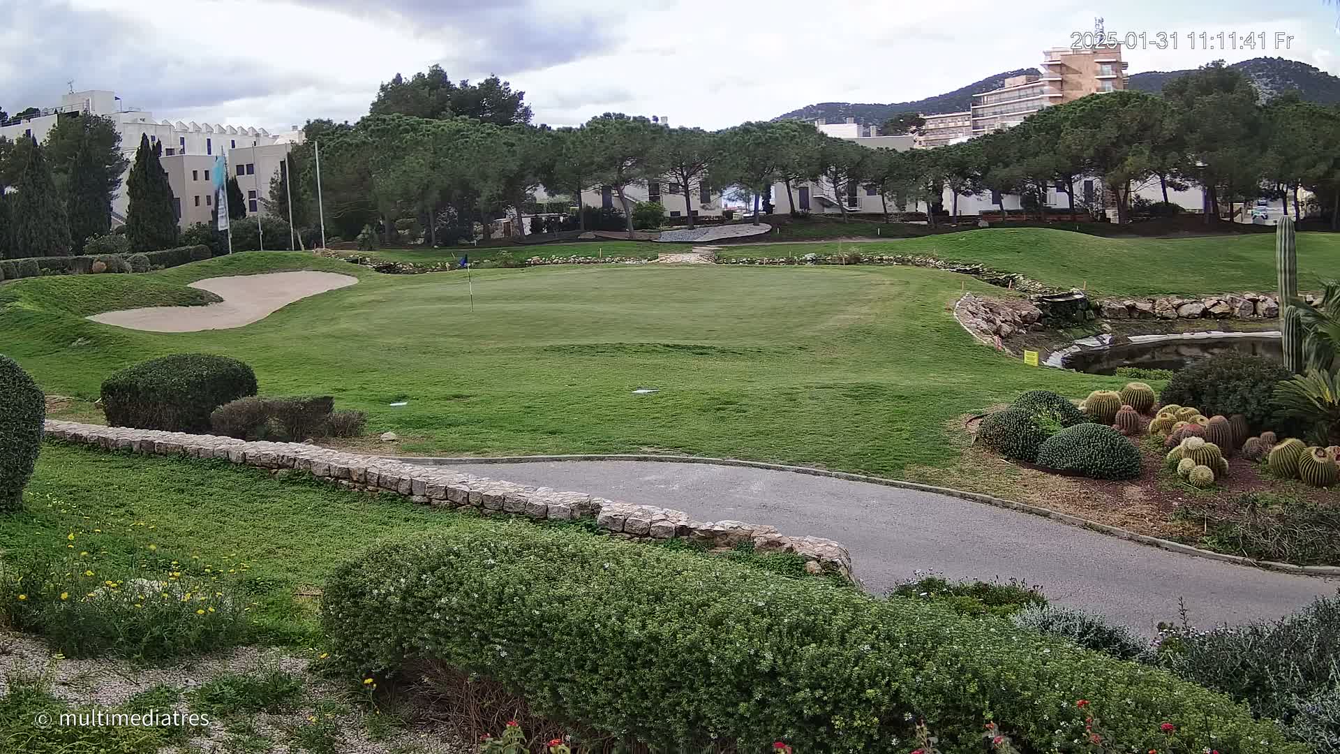 A golf course green with a sand trap, bordered by a low stone wall and landscaping including cacti and shrubs, under a cloudy sky.
