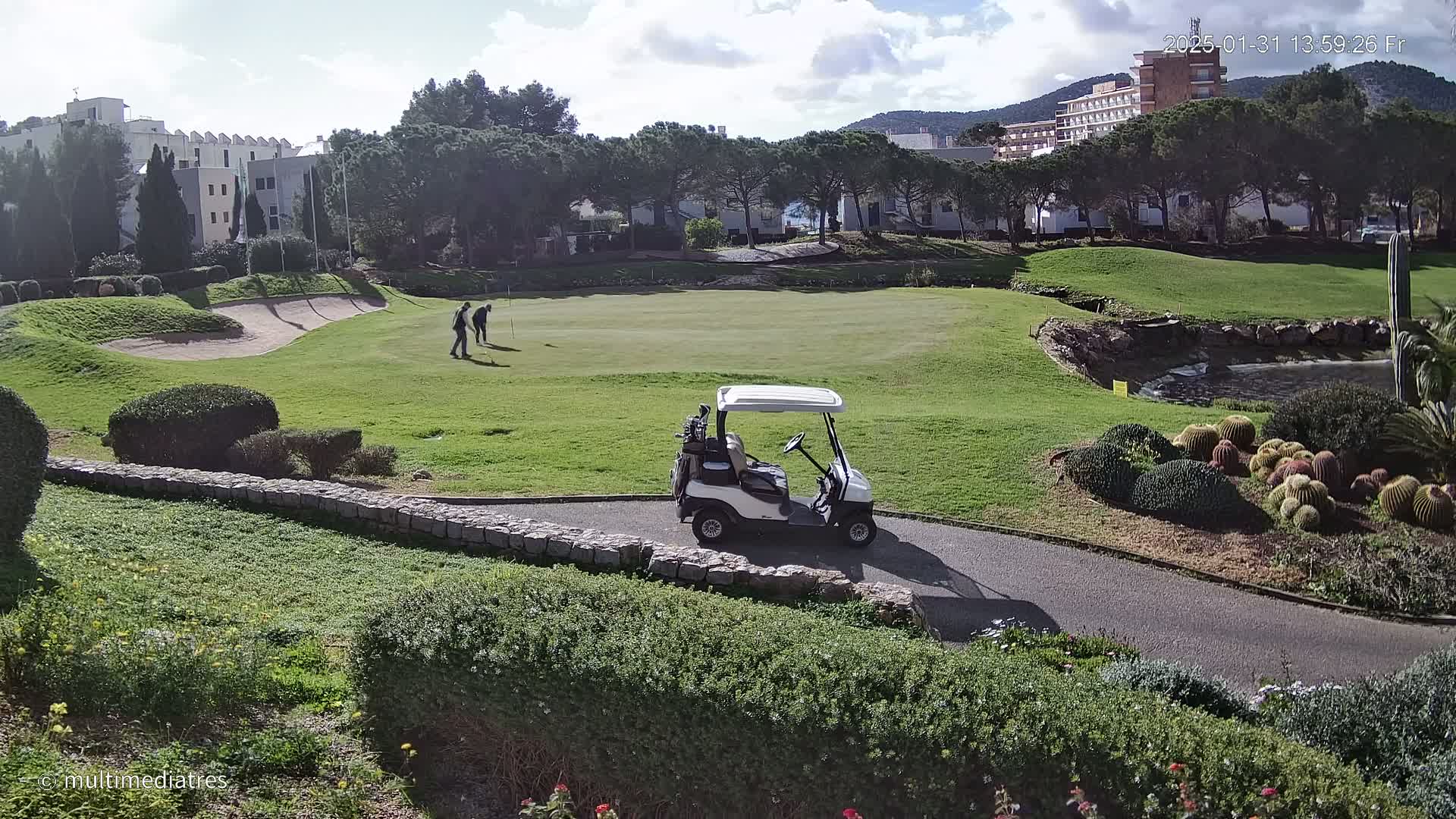 A golf course on a sunny day, with two golfers on the green and a golf cart parked nearby, surrounded by landscaping and buildings in the background.