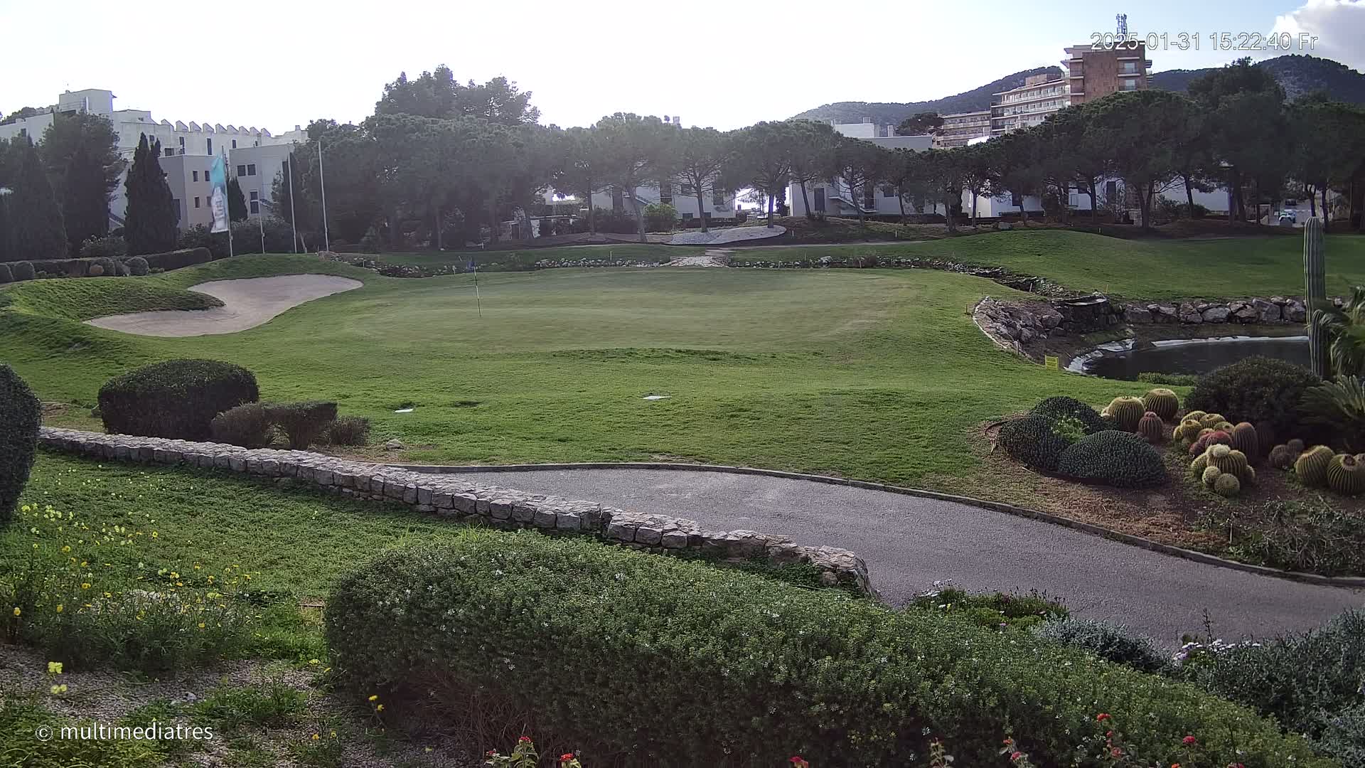 A golf course green with a sand trap, a small pond, and various shrubs and cacti, under a mostly cloudy sky.