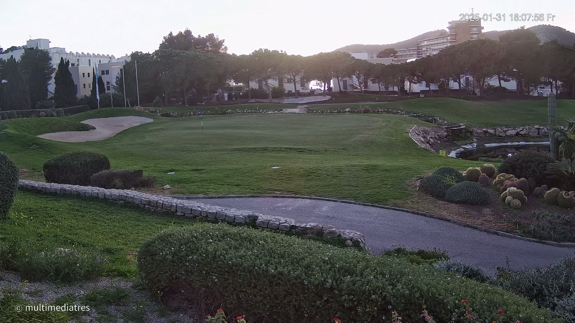 A golf course green with sand traps and a small pond, bordered by shrubs, a stone wall, and buildings in the background under a clear, twilight sky.