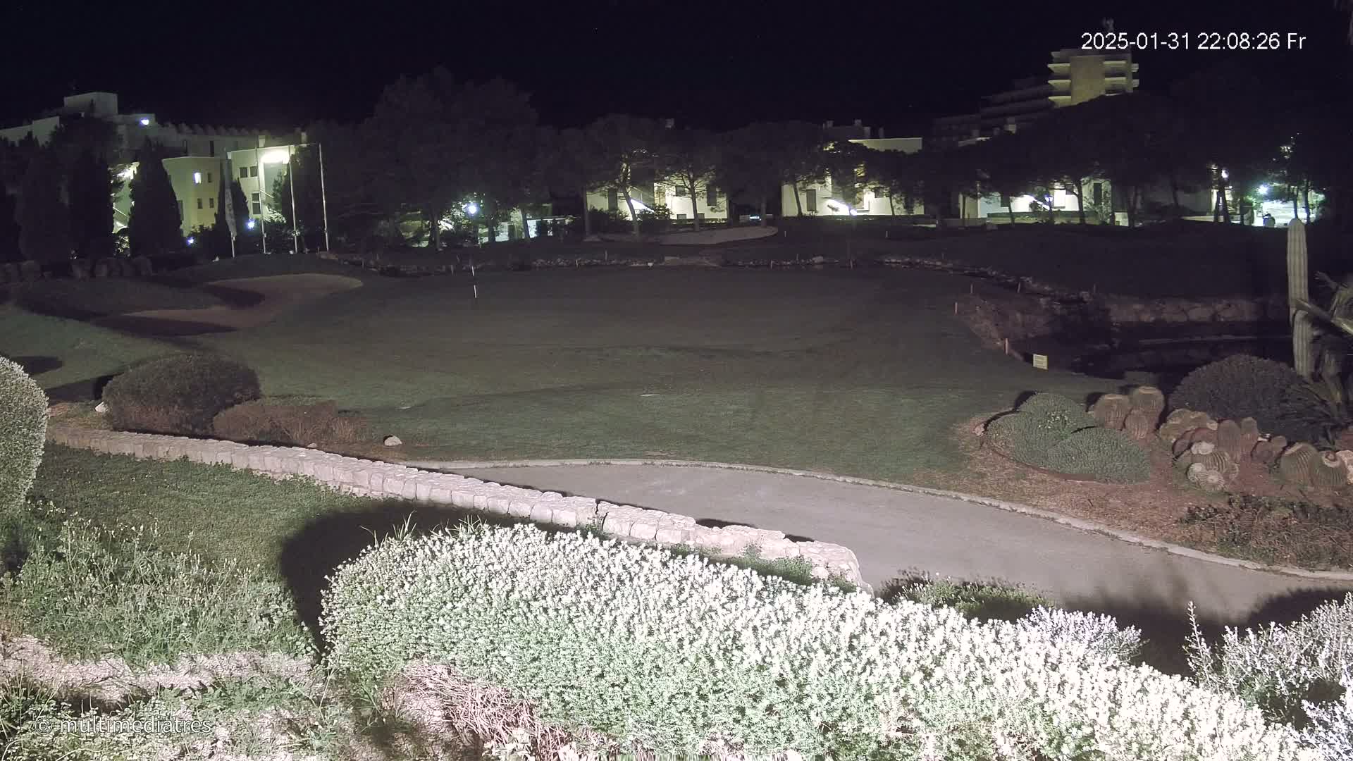 A nighttime view of a golf course, illuminated, with buildings in the background.