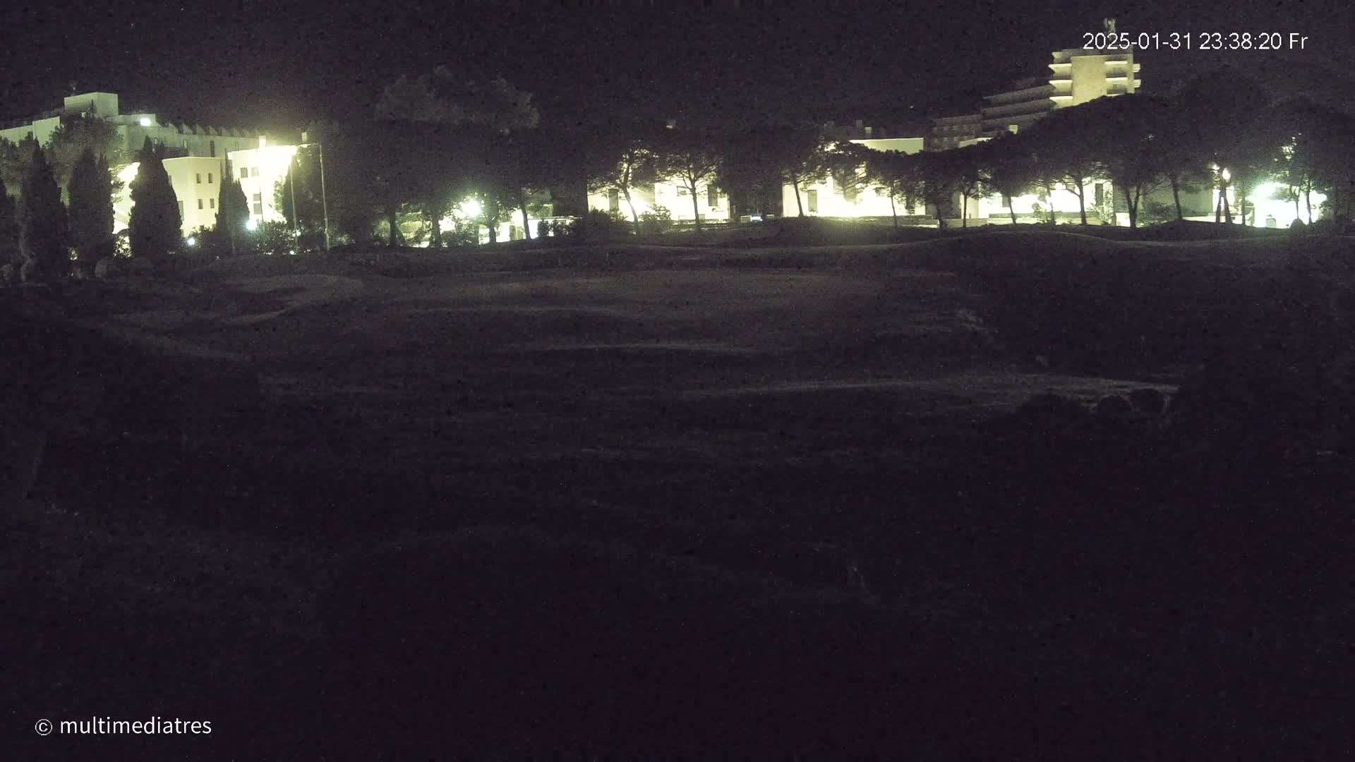 A nighttime view of a dark, grassy area with illuminated buildings and trees in the background.