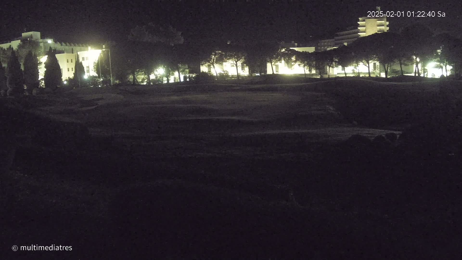 At night, illuminated buildings stand behind a dark, grassy area.