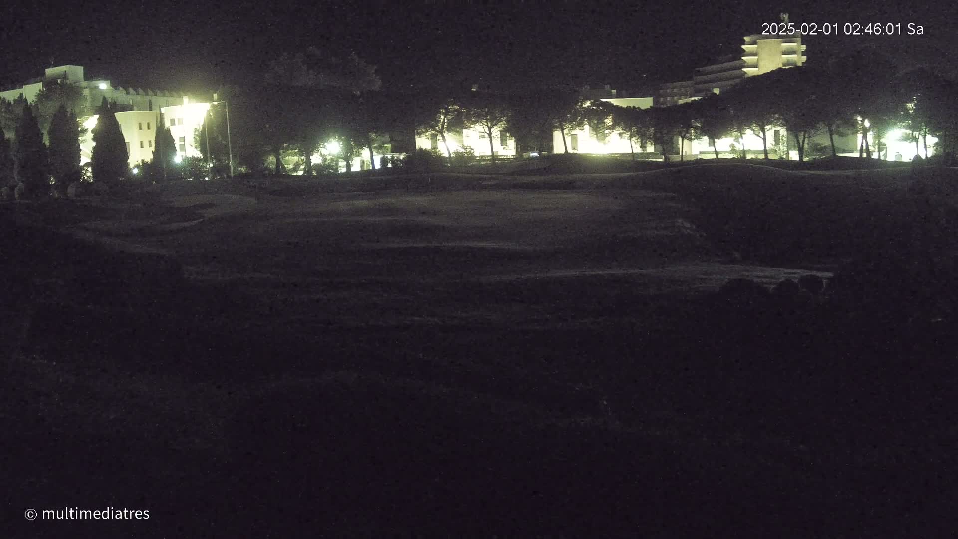 At night, a dark grassy area sits in front of illuminated buildings and trees.