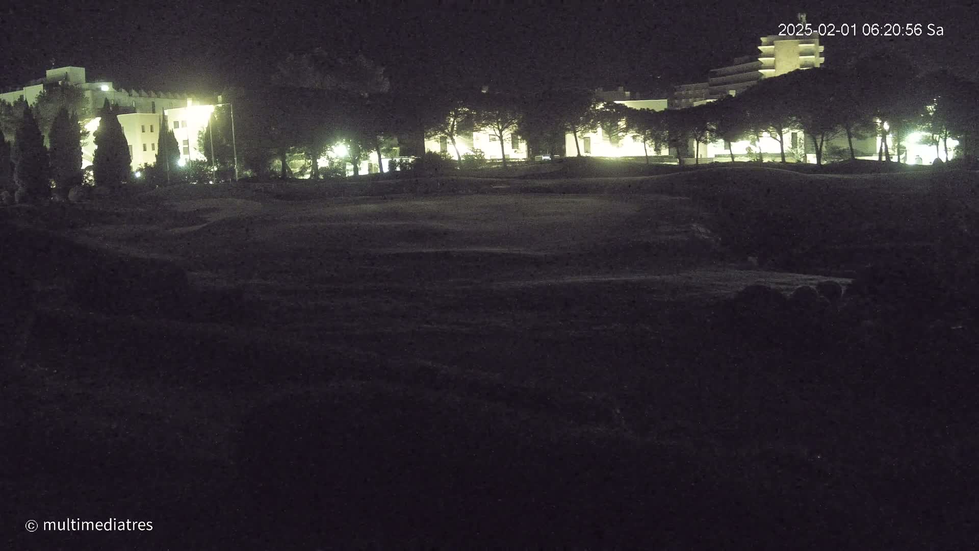 At night, illuminated buildings are visible behind a dark, grassy area.