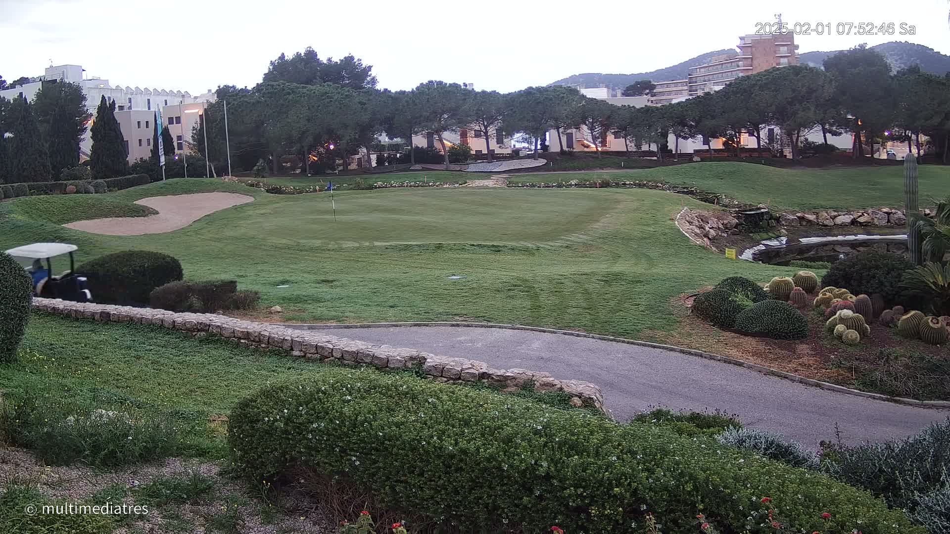 A golf course green with a sand trap, a golf cart, and a low stone wall is visible, with a hotel and pine trees in the background under an overcast sky.