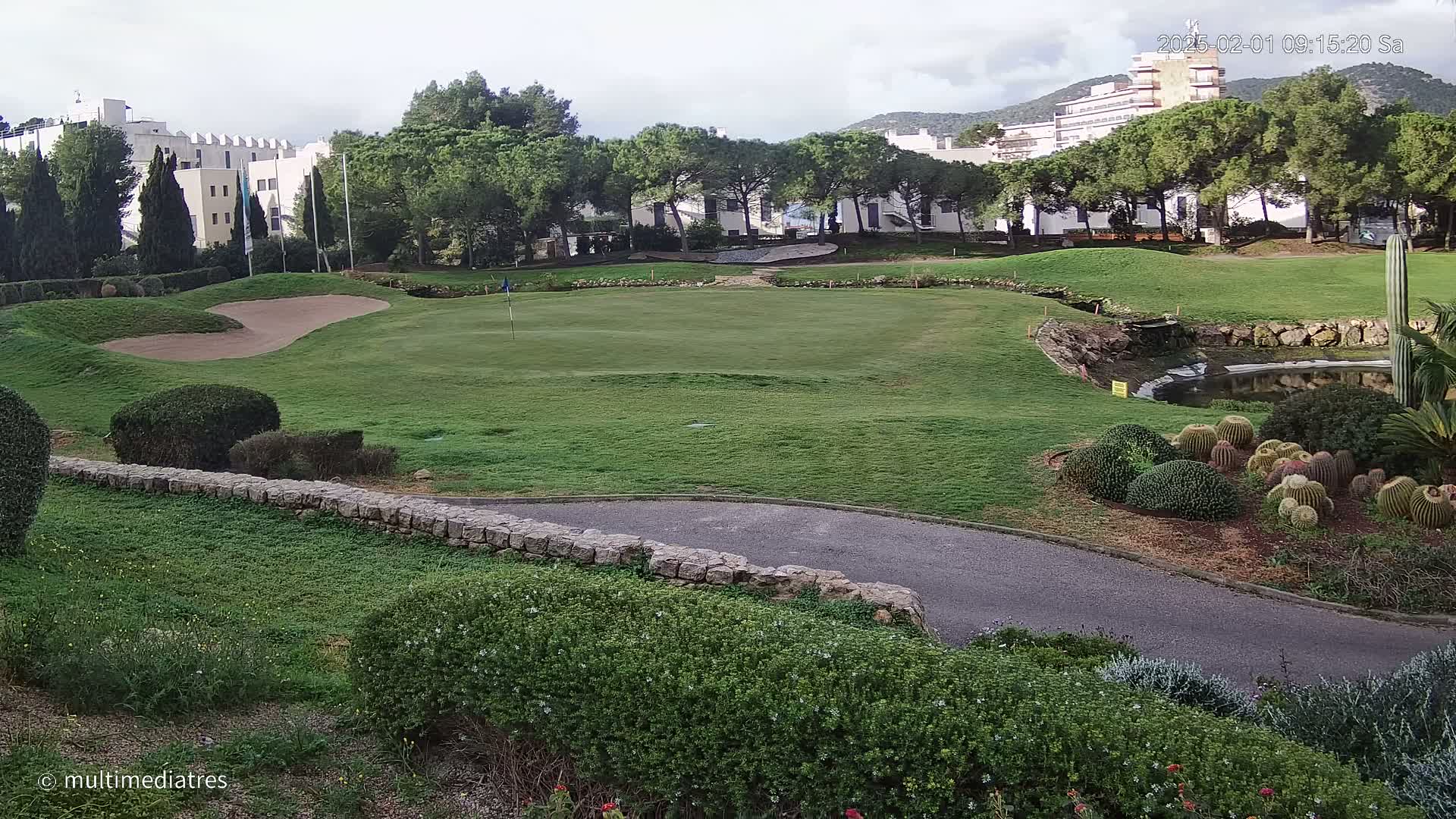 A golf course green with a sand trap, bordered by shrubs, a low stone wall, and a paved path, situated near buildings and under a partly cloudy sky.
