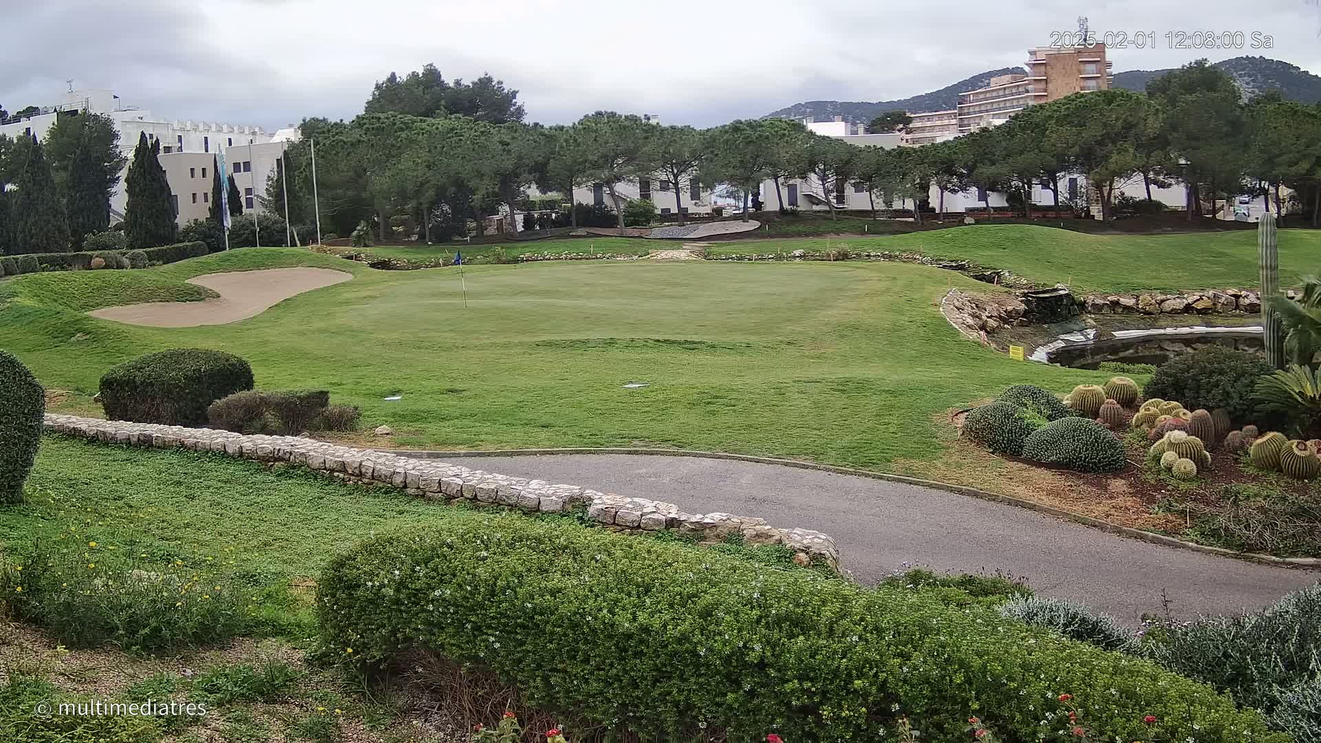 A golf course green with sand trap and a small pond is seen on an overcast day, next to a low stone wall, with buildings and trees in the background.