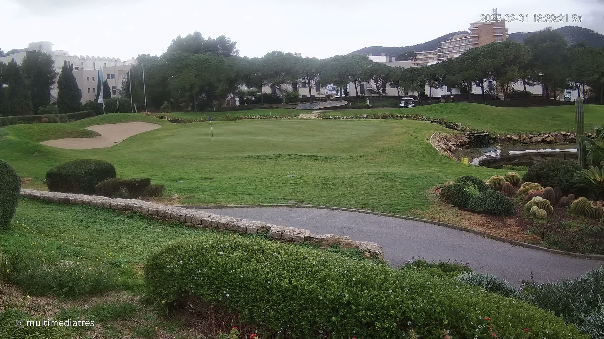 A golf course green with a sand trap and a small pond, situated near buildings and trees under an overcast sky.