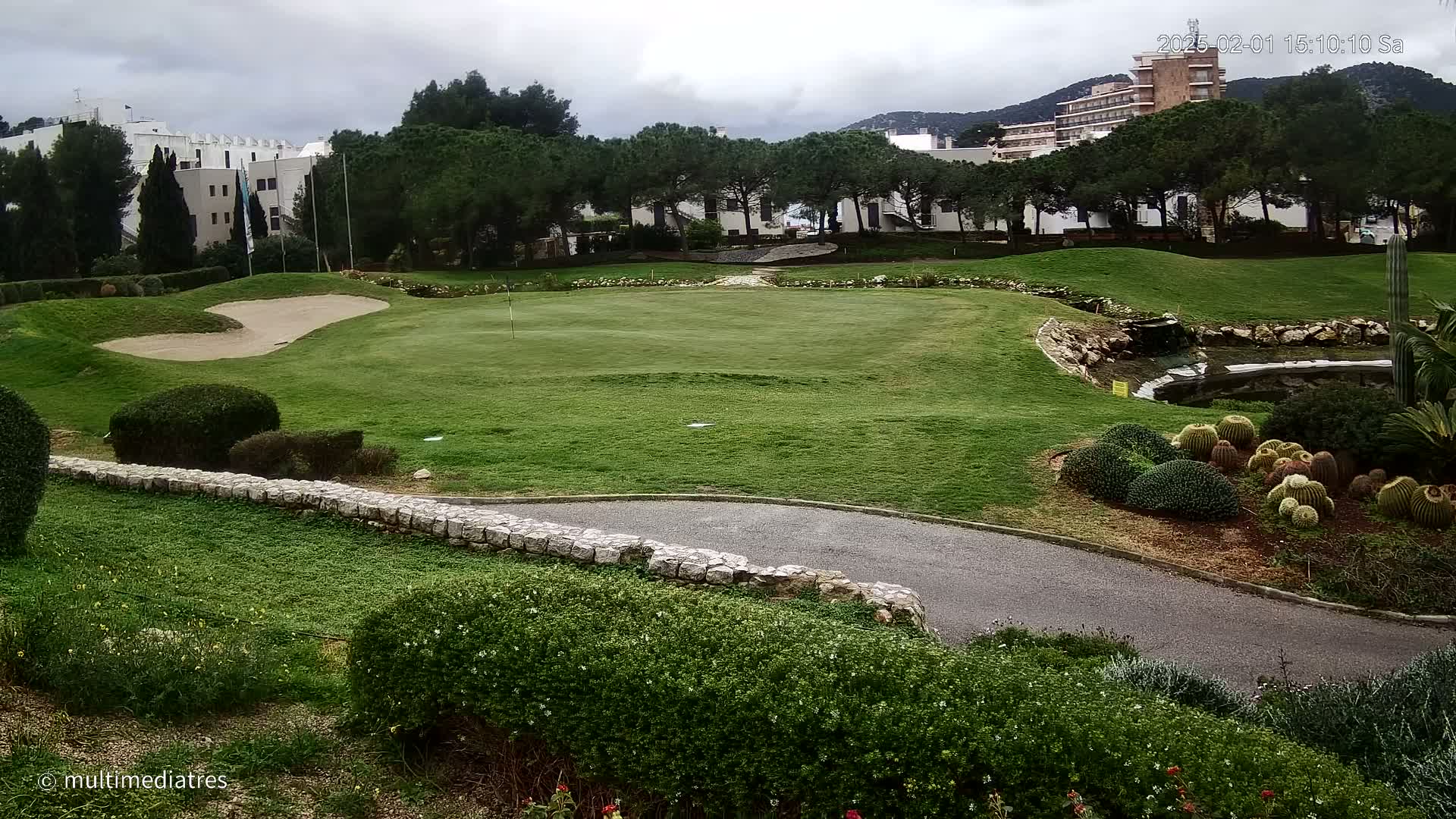 A golf course green with a sand trap, bordered by manicured shrubs and a low stone wall, overlooks a small pond and buildings under a cloudy sky.