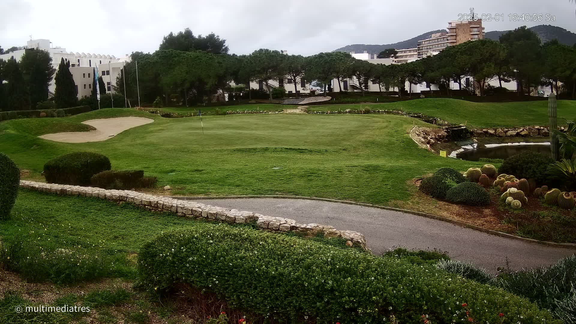 A golf course green with a sand trap, a small pond, and manicured landscaping is situated in front of a building and some trees under an overcast sky.