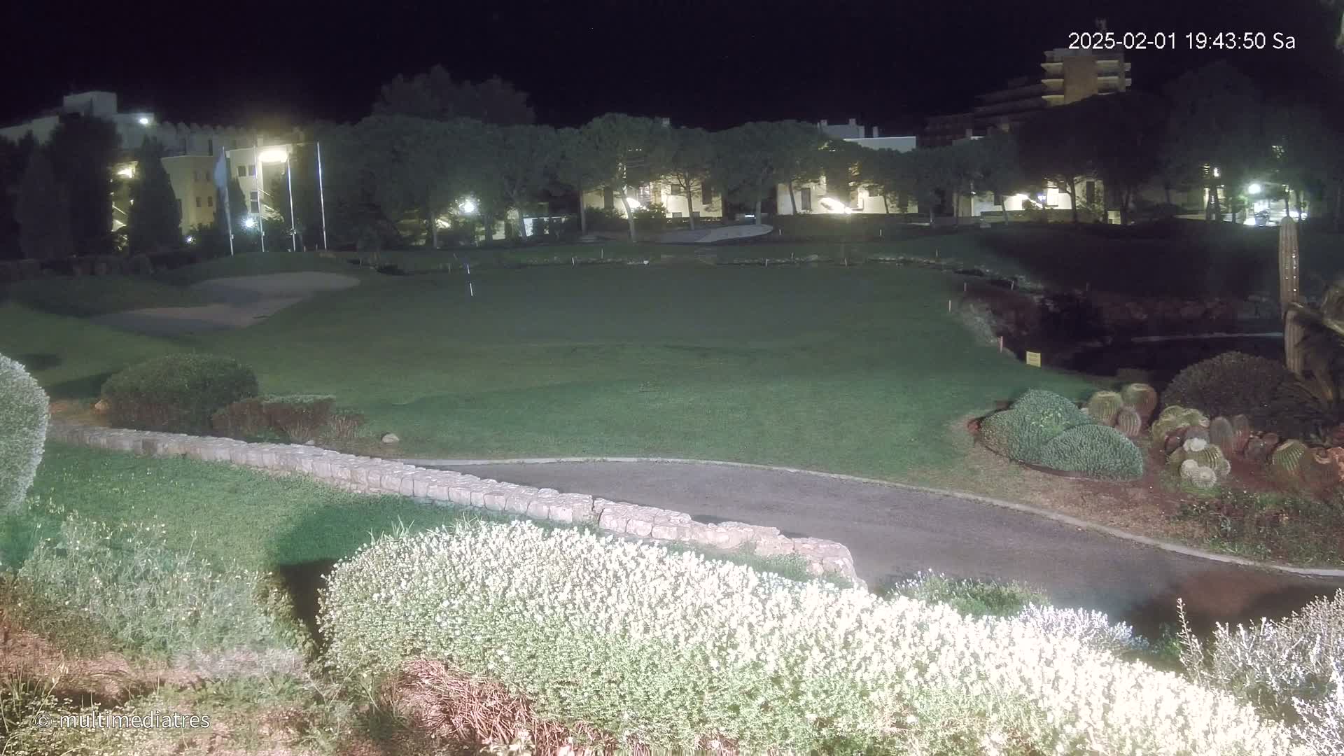 A nighttime view of a golf course putting green, bordered by low stone walls and shrubs, with a building in the background under artificial light.